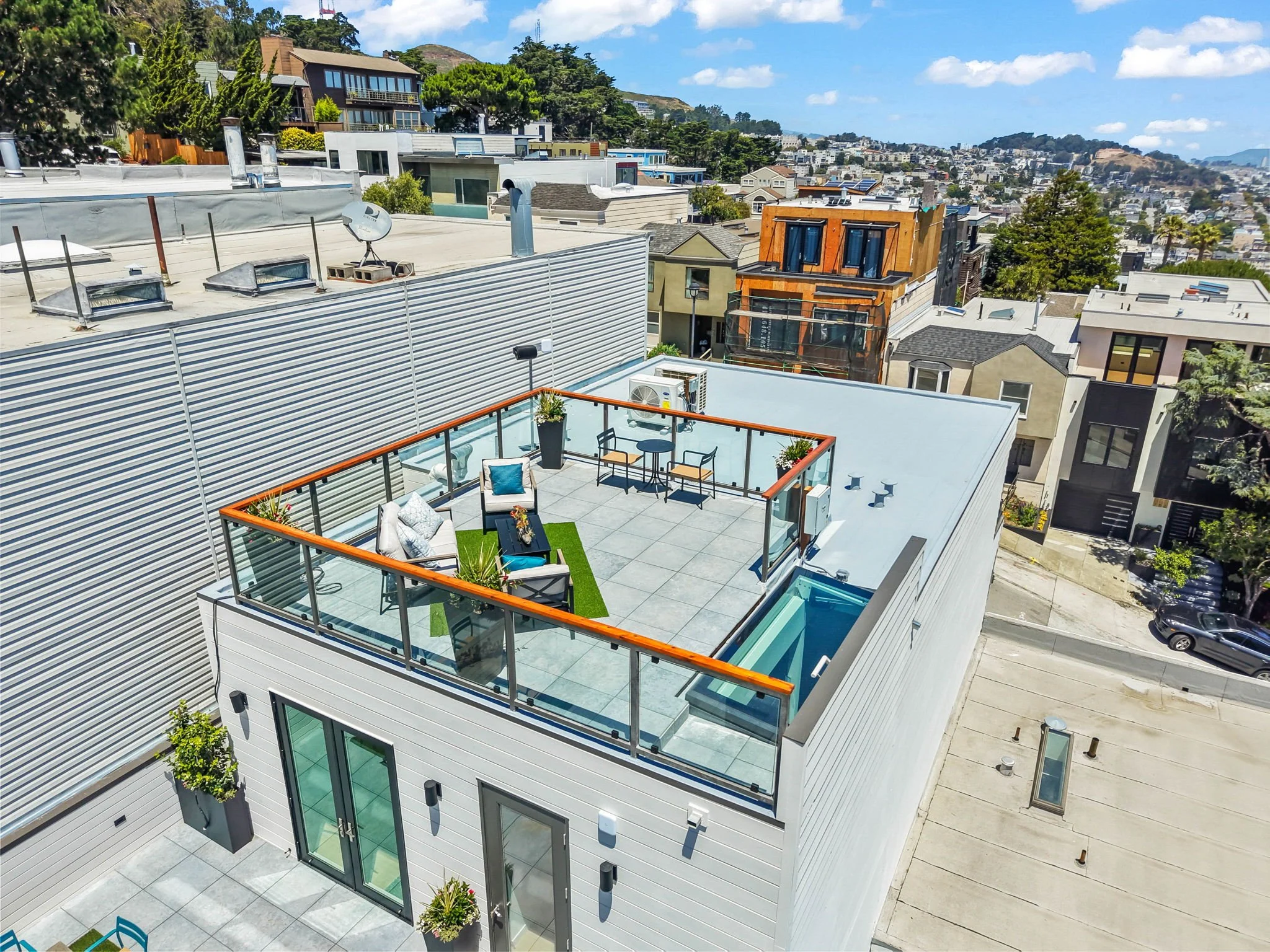 A view of a modern rooftop patio with outdoor furniture, potted plants, and a glass railing, overlooking a neighborhood with various residential buildings and trees under a partly cloudy sky.