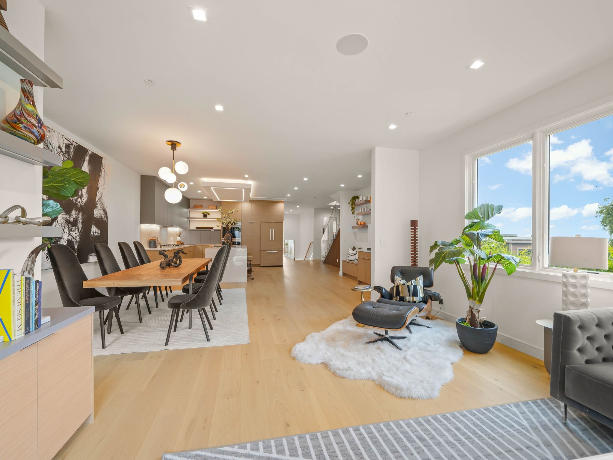 Modern open-concept kitchen and living room with large windows, wooden floor, chandelier, potted plants, dining area, and seating furnishings.