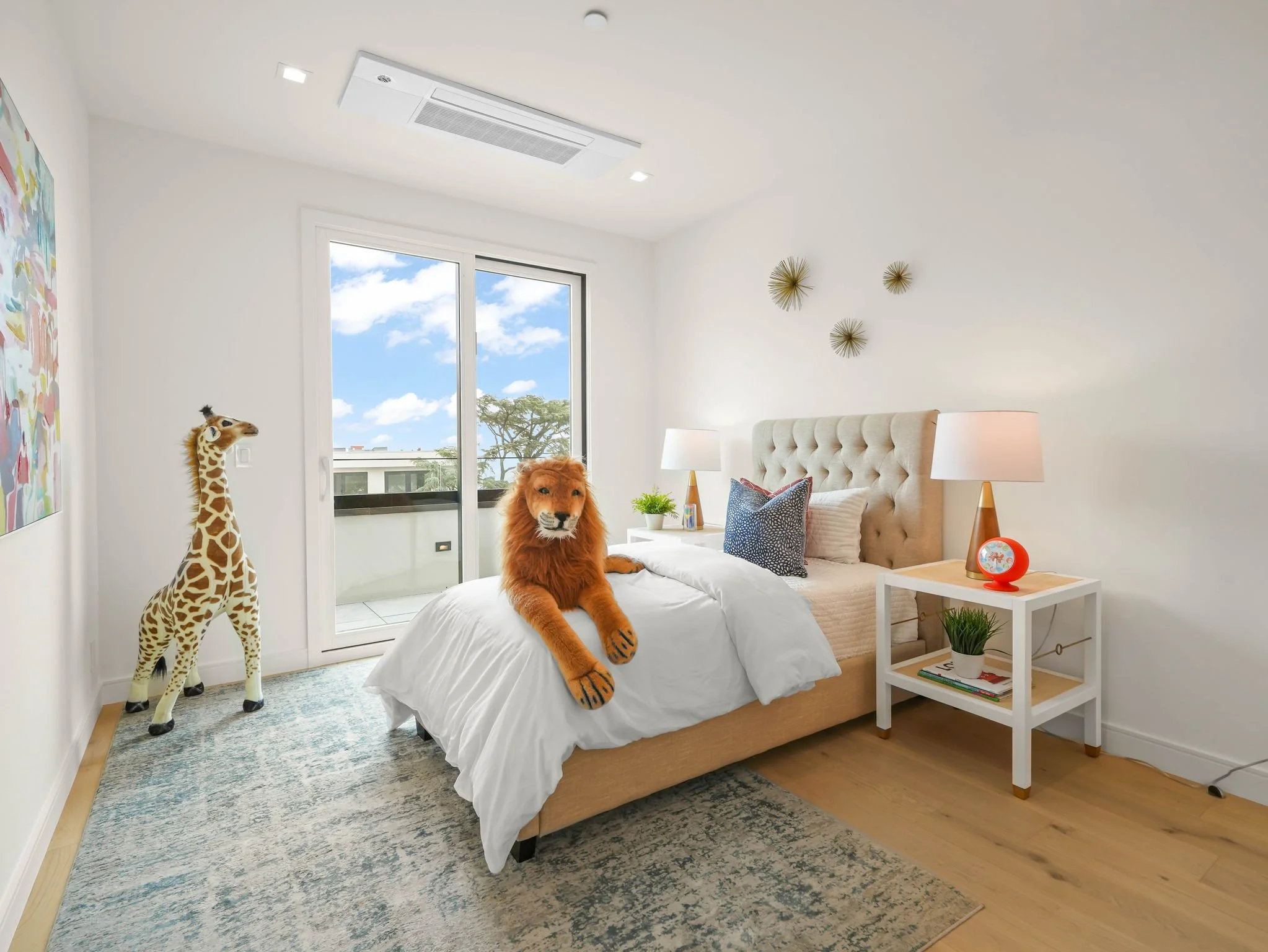 A bedroom with white walls and a large window showing blue sky and trees outside. There is a beige upholstered bed with a white duvet and pillows, including a blue patterned pillow. On the bed, there is a realistic stuffed lion and a giraffe. Next to