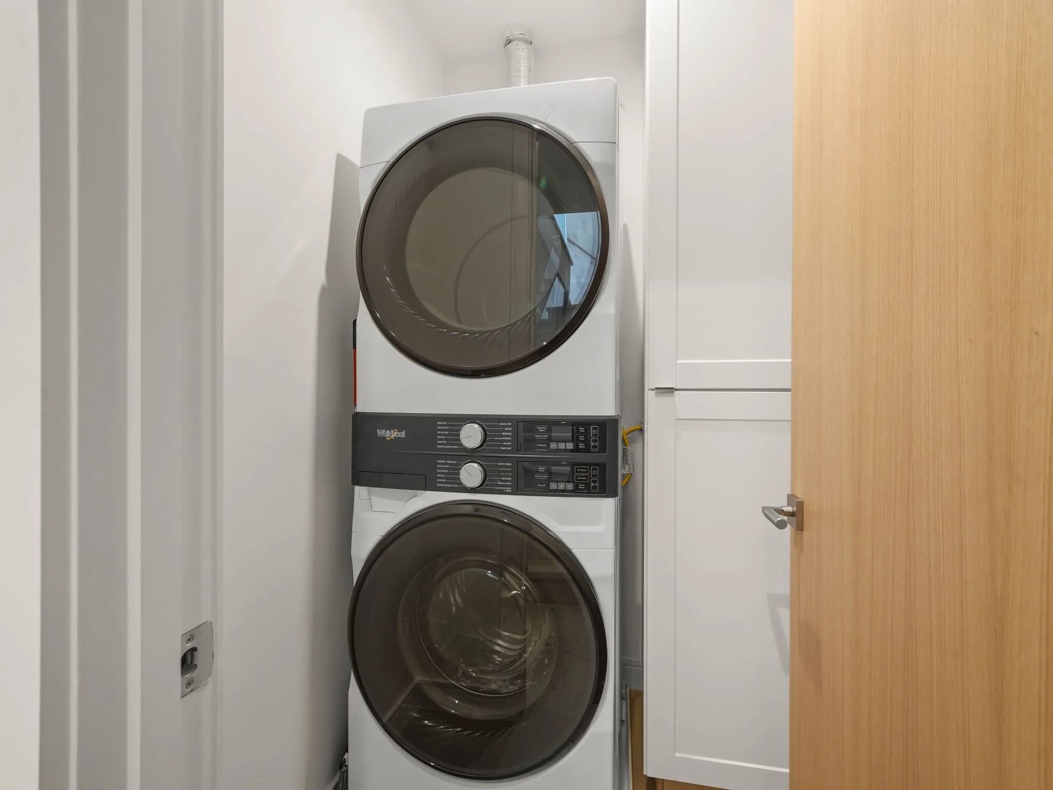 Stacked white washing machine and dryer in a laundry room with wood floor and white walls.