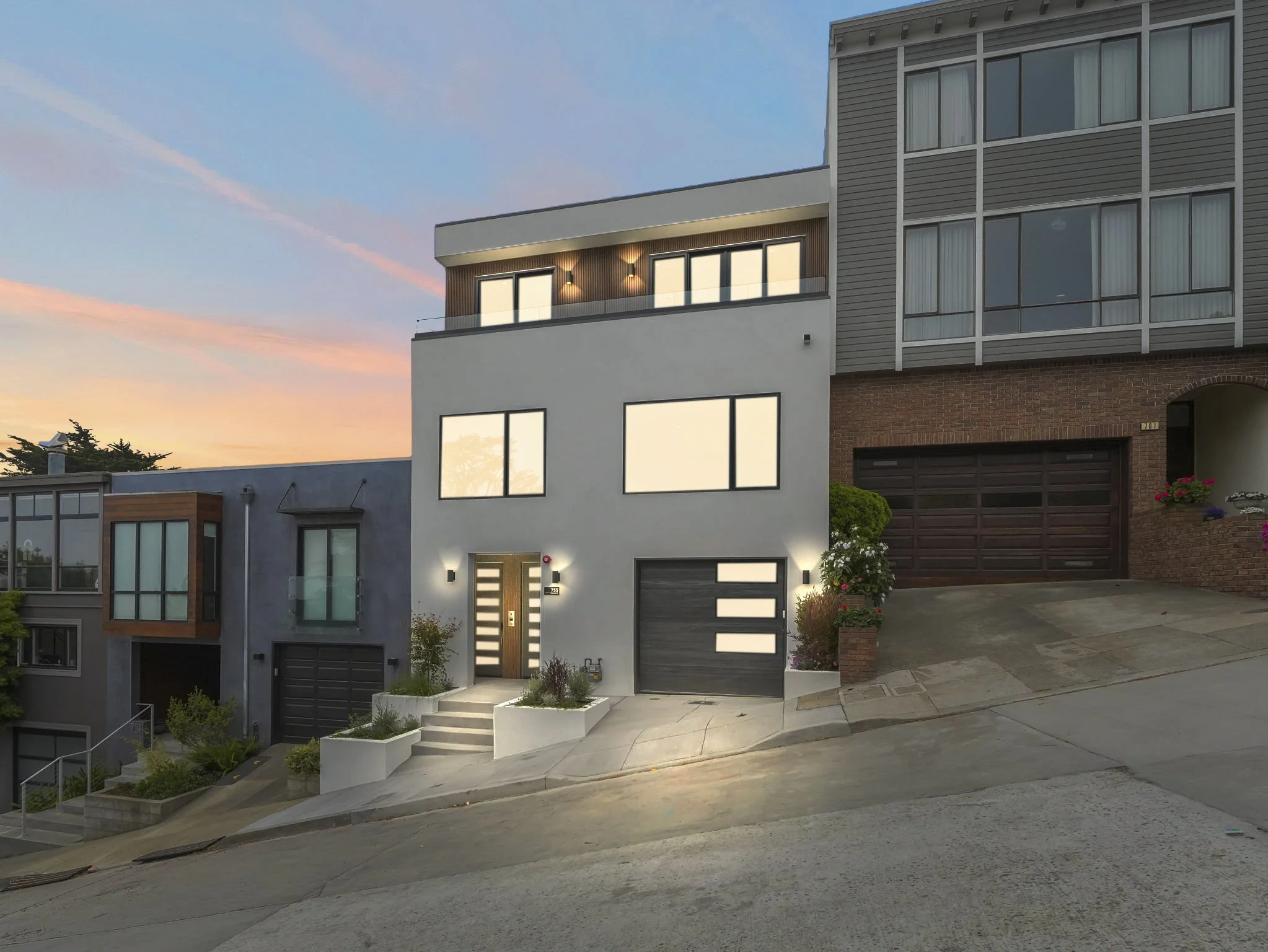 Modern multi-story residential building with large windows, a combination of brick, wood, and siding exterior, front steps, and small landscaped area with plants at the entrance. Sunset sky in the background.