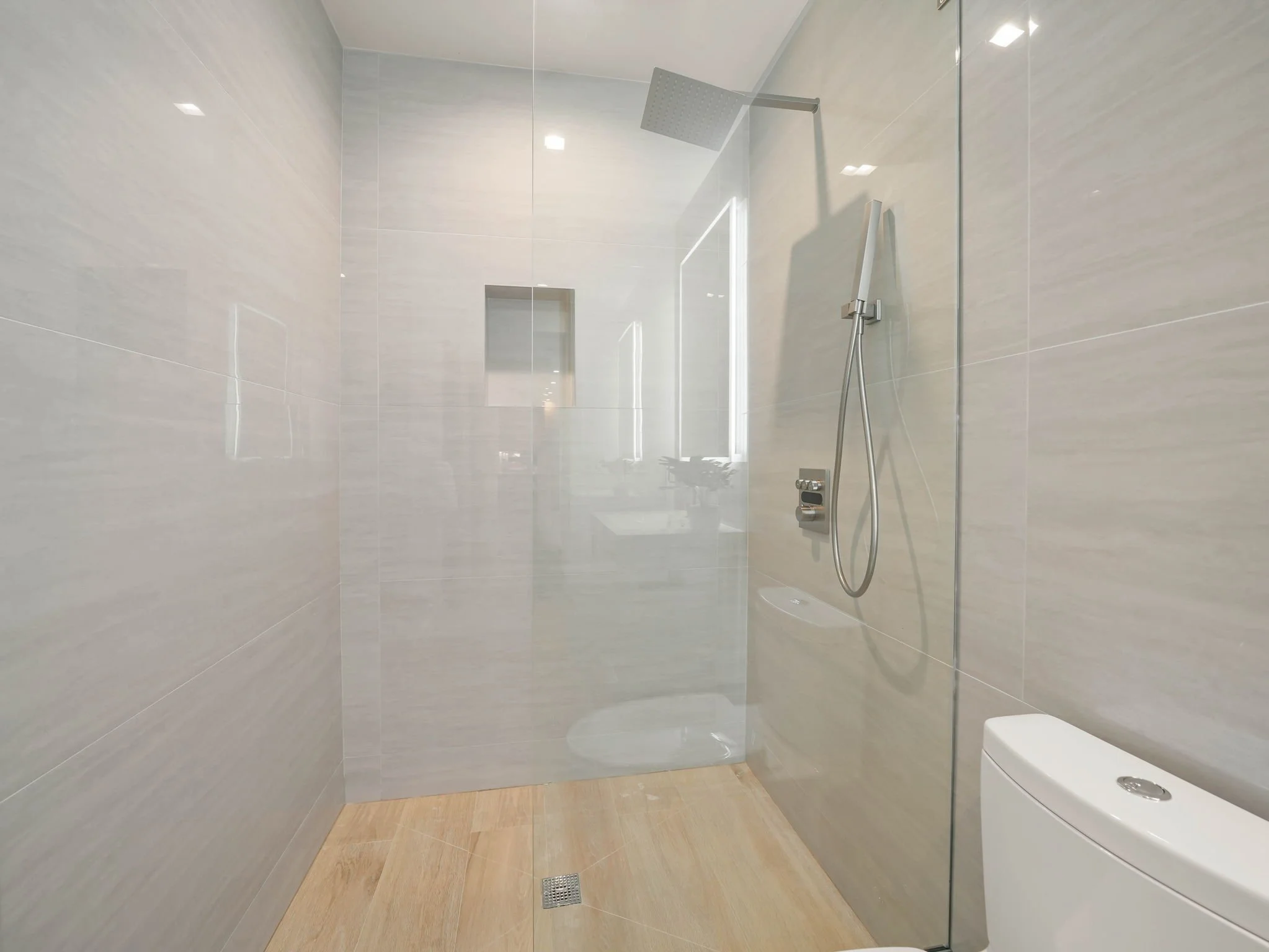 Modern walk-in shower with large square rain shower head, handheld showerhead, beige tiles on the floor, and large light-colored wall tiles, with a small wall-mounted niche and a glass partition.