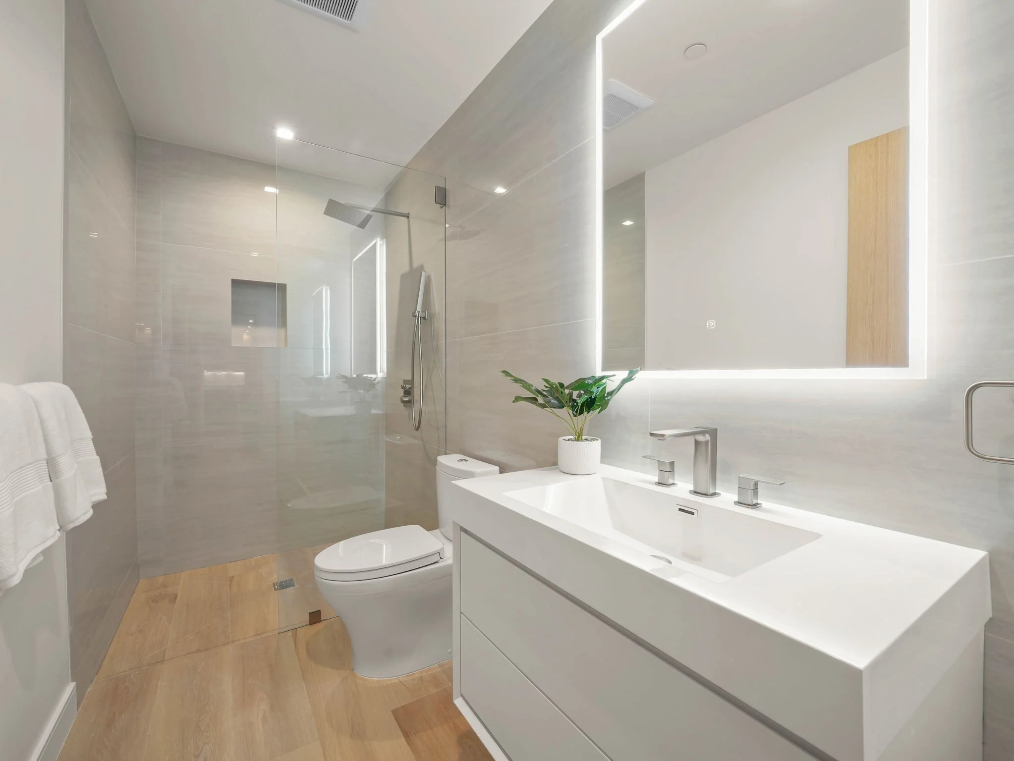 Modern bathroom with a wall-mounted toilet, white vanity with dual sinks, large mirror with LED lighting, and a walk-in shower with glass enclosure and overhead rainfall showerhead. There is a potted plant on the vanity and wooden flooring.