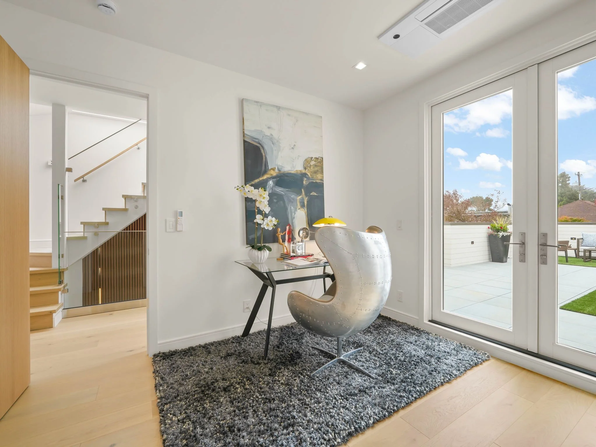 Interior view of a bright room with large glass doors leading outside, a modern chair, a small black table, a gray shag rug, a large abstract painting on the wall, and decorative items on the table, with a view of the backyard.