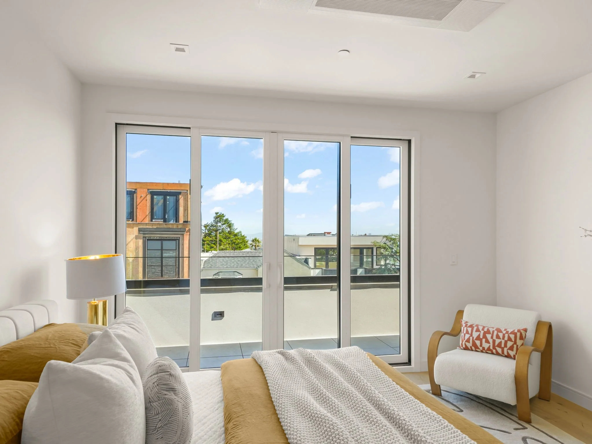 Living room with white walls and a large glass sliding door opening to a balcony with a view of a building, trees, and blue sky with clouds. There is a beige couch with white and brown pillows, and a gold-colored lamp on a side table. A modern wooden