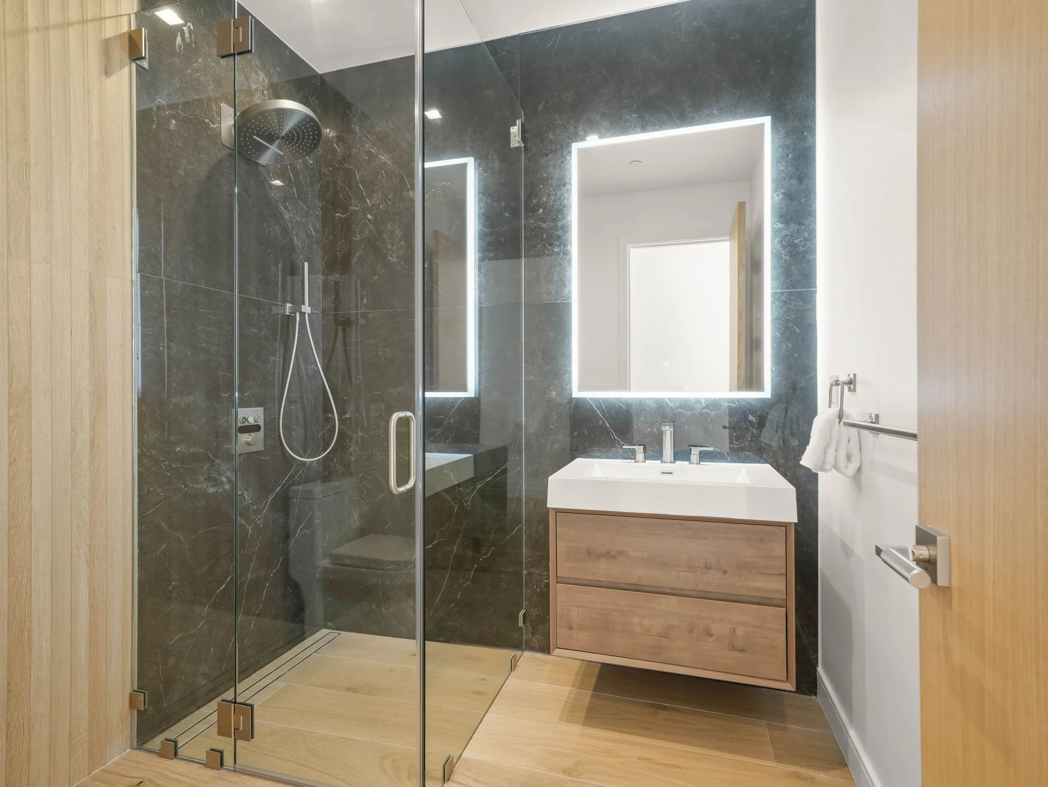 Modern bathroom with a glass shower enclosure, marble walls, wooden vanity with a sink, illuminated mirror, and wooden flooring.