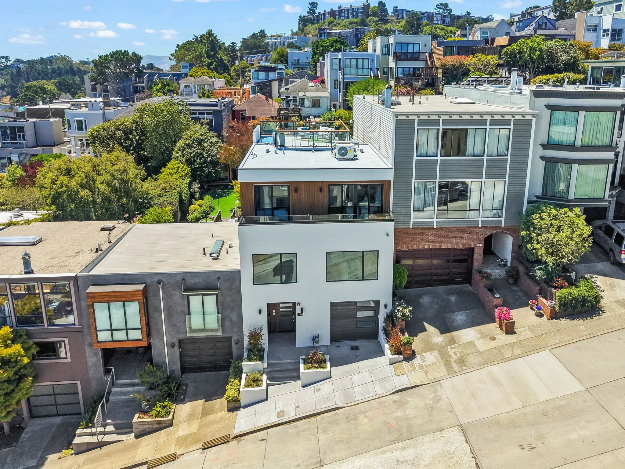 Aerial view of a neighborhood with modern multi-story houses on a hillside, lush trees, and a wide sidewalk in the foreground on a sunny day.