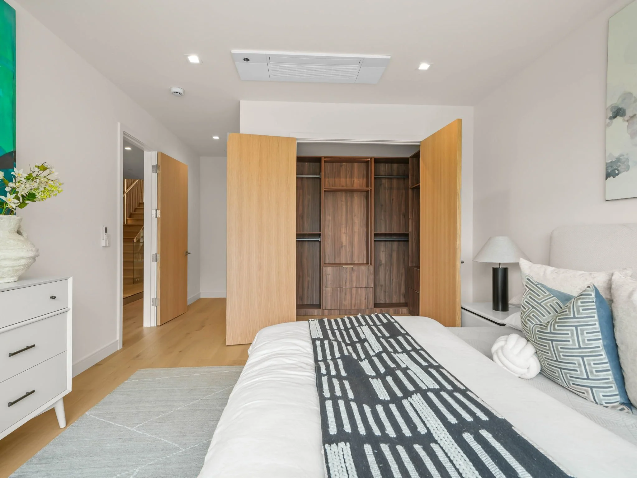 A bedroom with a bed, pillows, and a black and white striped runner, a white bedside table with a lamp, a vase of flowers, an open wooden closet, a lamp, a painting, and a doorway leading upstairs.