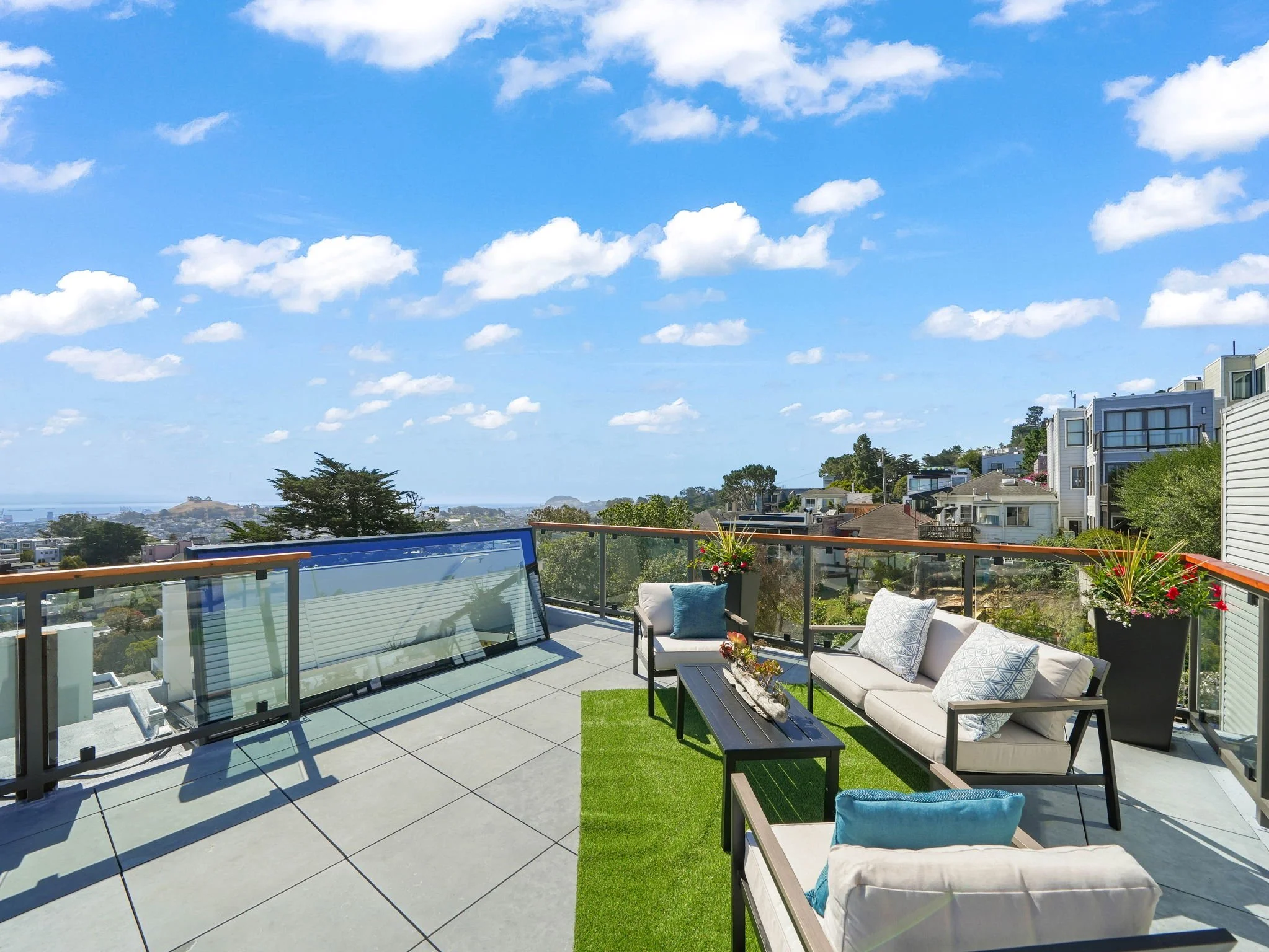 View of a rooftop patio with outdoor furniture, including chairs and a table, overlooking a coastal neighborhood with houses and a hill in the background under a blue sky with scattered clouds.