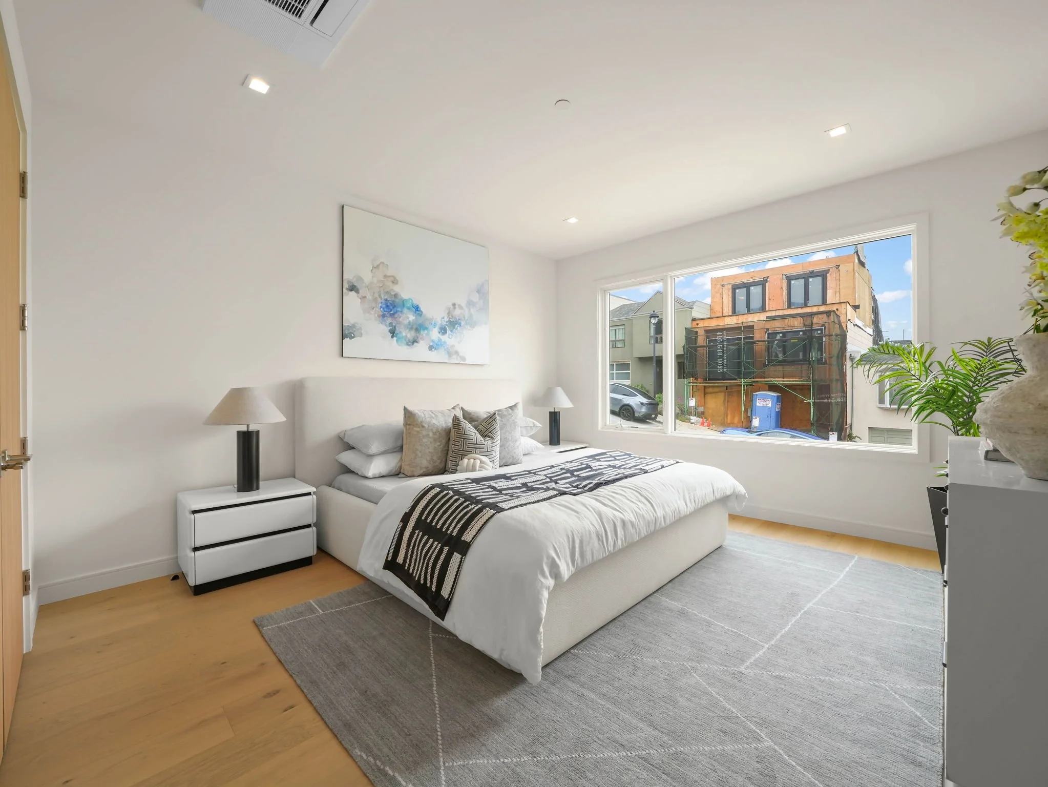 A bedroom with a white bed, black and white bedding, and pillows. There are two matching nightstands with lamps, and a large window showing a street scene with a building under construction.