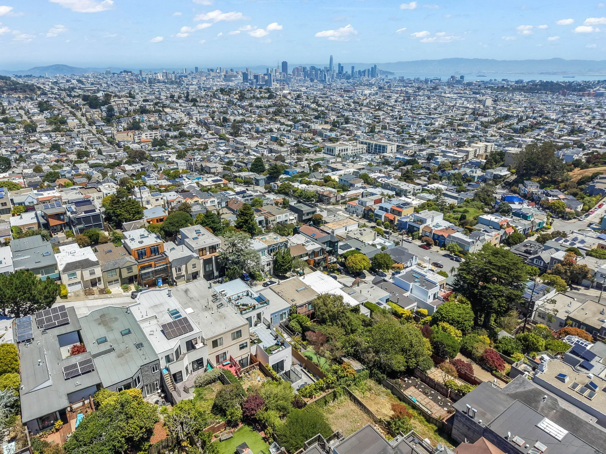Aerial view of a densely populated cityscape with a mix of residential houses and tall skyscrapers against a backdrop of water and hills under a partly cloudy sky.