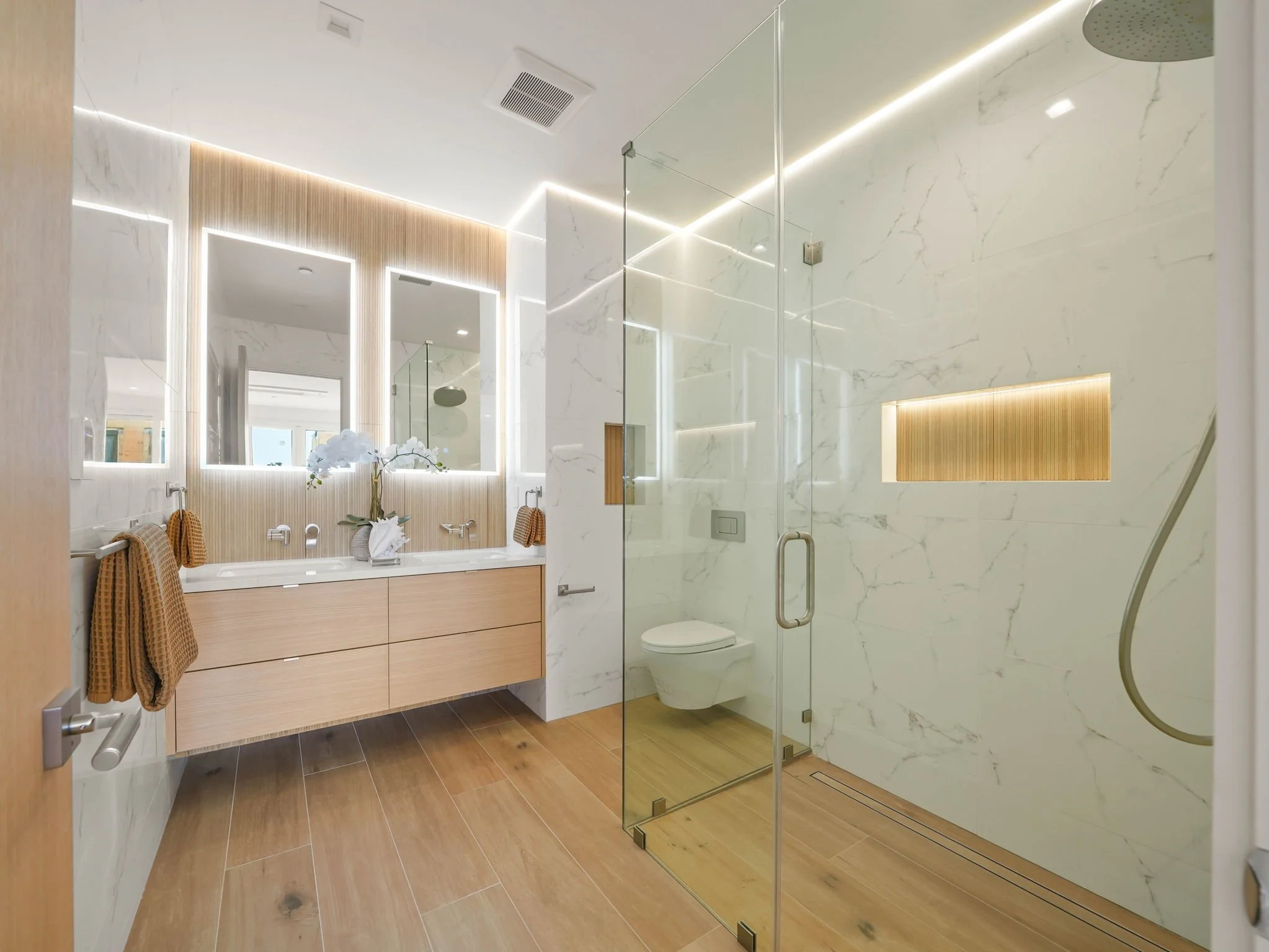 Modern bathroom with marble walls and wooden flooring, featuring a glass shower enclosure, a floating vanity with a large mirror, and a toilet. Decorative towels, a vase with white flowers, and warm lighting accents are visible.