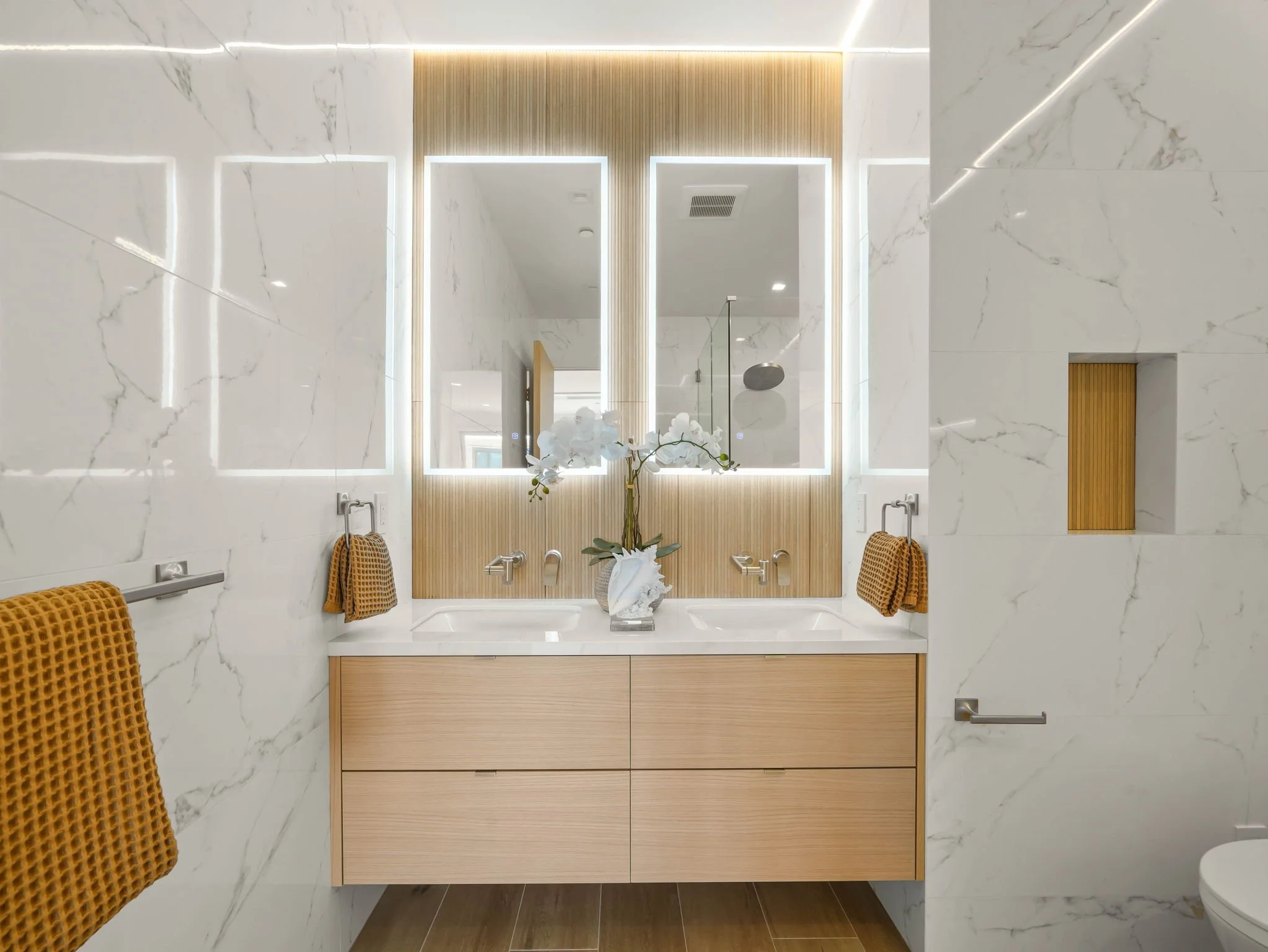 Modern bathroom with marble walls, a wooden vanity with a sink, a large mirror above, and decorative white orchids.