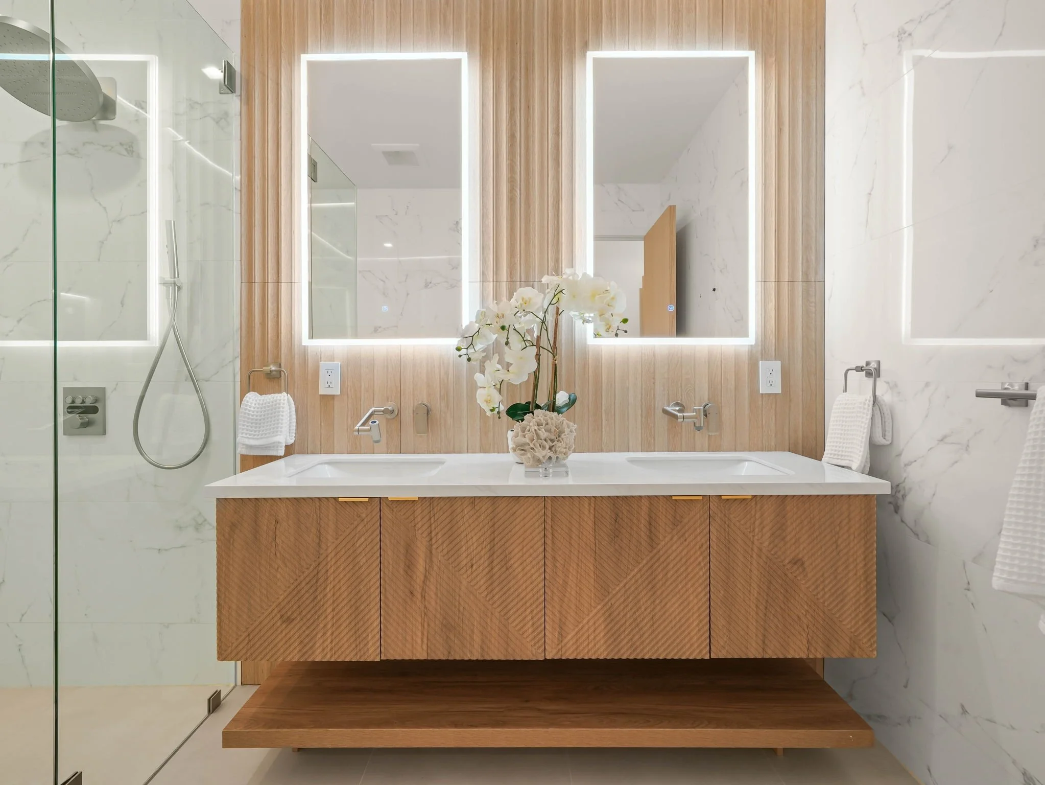 Modern bathroom with marble walls, a wooden vanity with a white vessel sink, a flower arrangement, mirrored medicine cabinets with built-in lighting, and a glass shower enclosure.