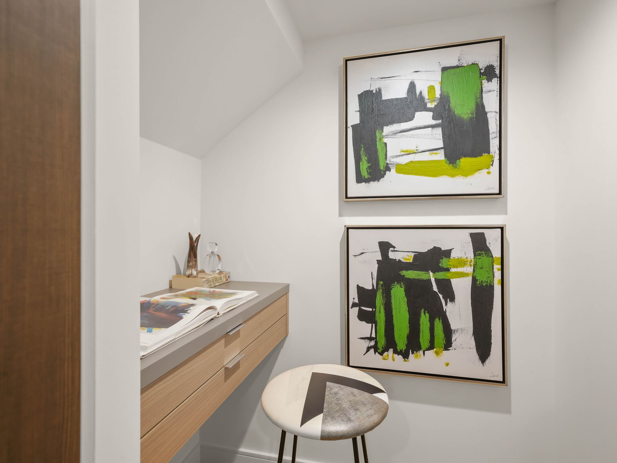 A modern interior space featuring abstract artwork with black, green, and gray brushstrokes on a white wall, a light wood sideboard with books and decorative objects, a small round table with a patterned chair, and a white wall with a curved ceiling.