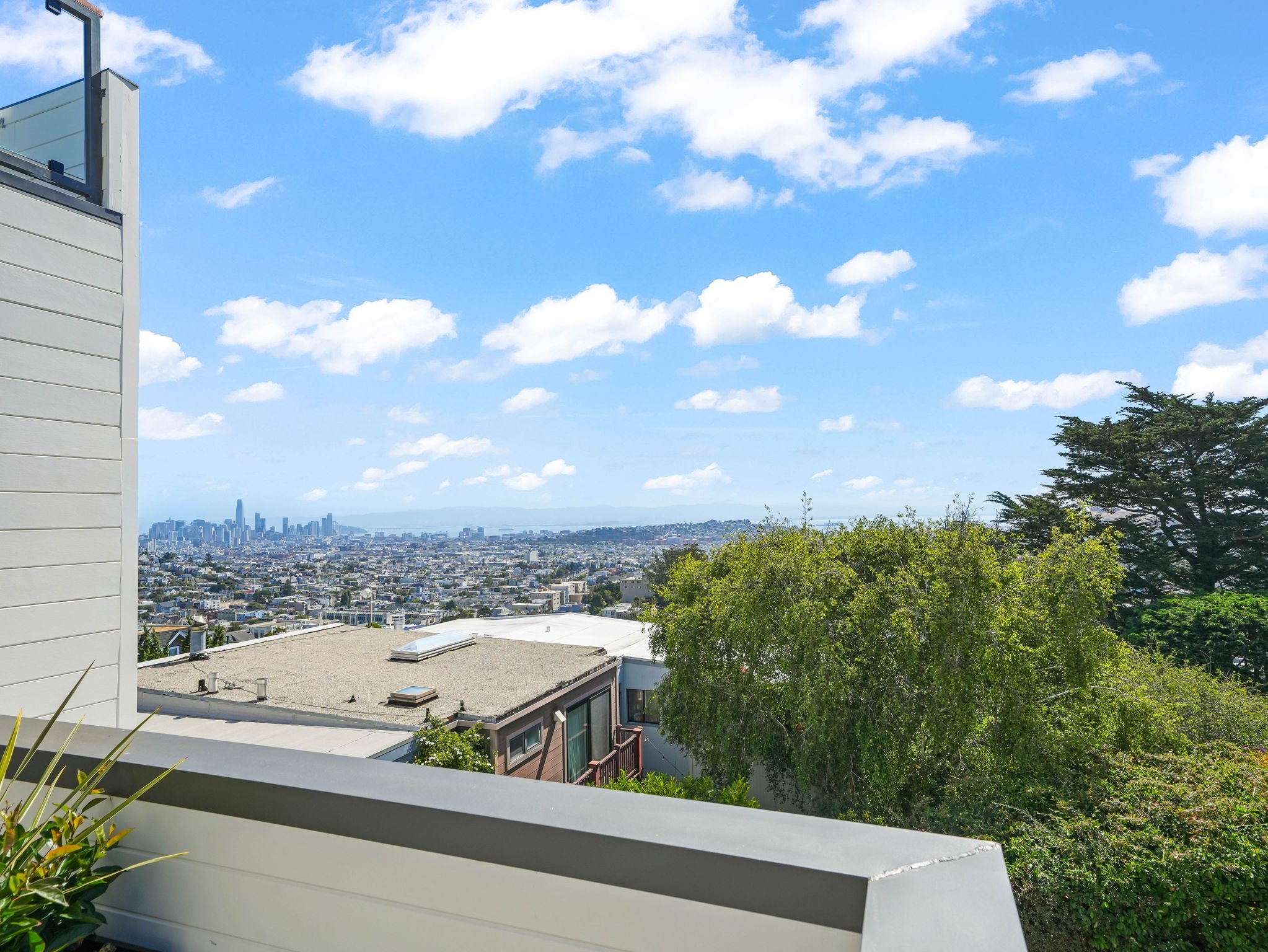 View of a city skyline in the distance with a clear blue sky and scattered clouds. The foreground shows residential buildings, trees, and part of a balcony or patio.