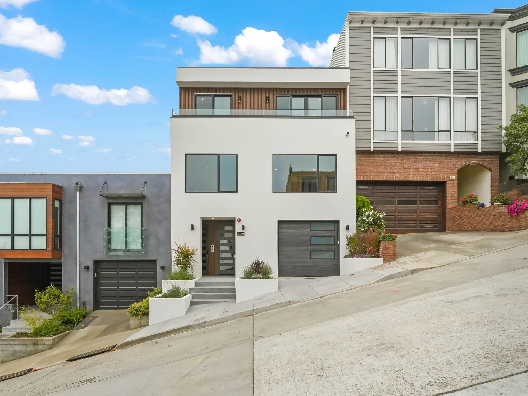 Front view of modern multi-story residential building with a variety of architectural styles, large windows, and a garage door. The house is situated on a sloped cement driveway with small landscaped areas and shrubs, under a partly cloudy blue sky.