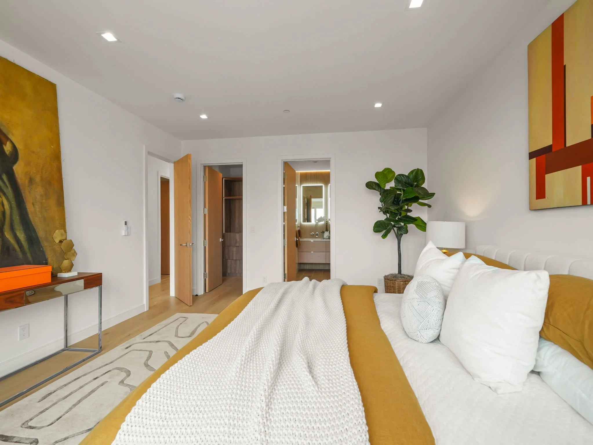 Modern bedroom with white walls and ceiling, wooden floors, a bed with white and mustard-colored bedding, a large potted plant, abstract artwork, and open doors leading to other rooms.