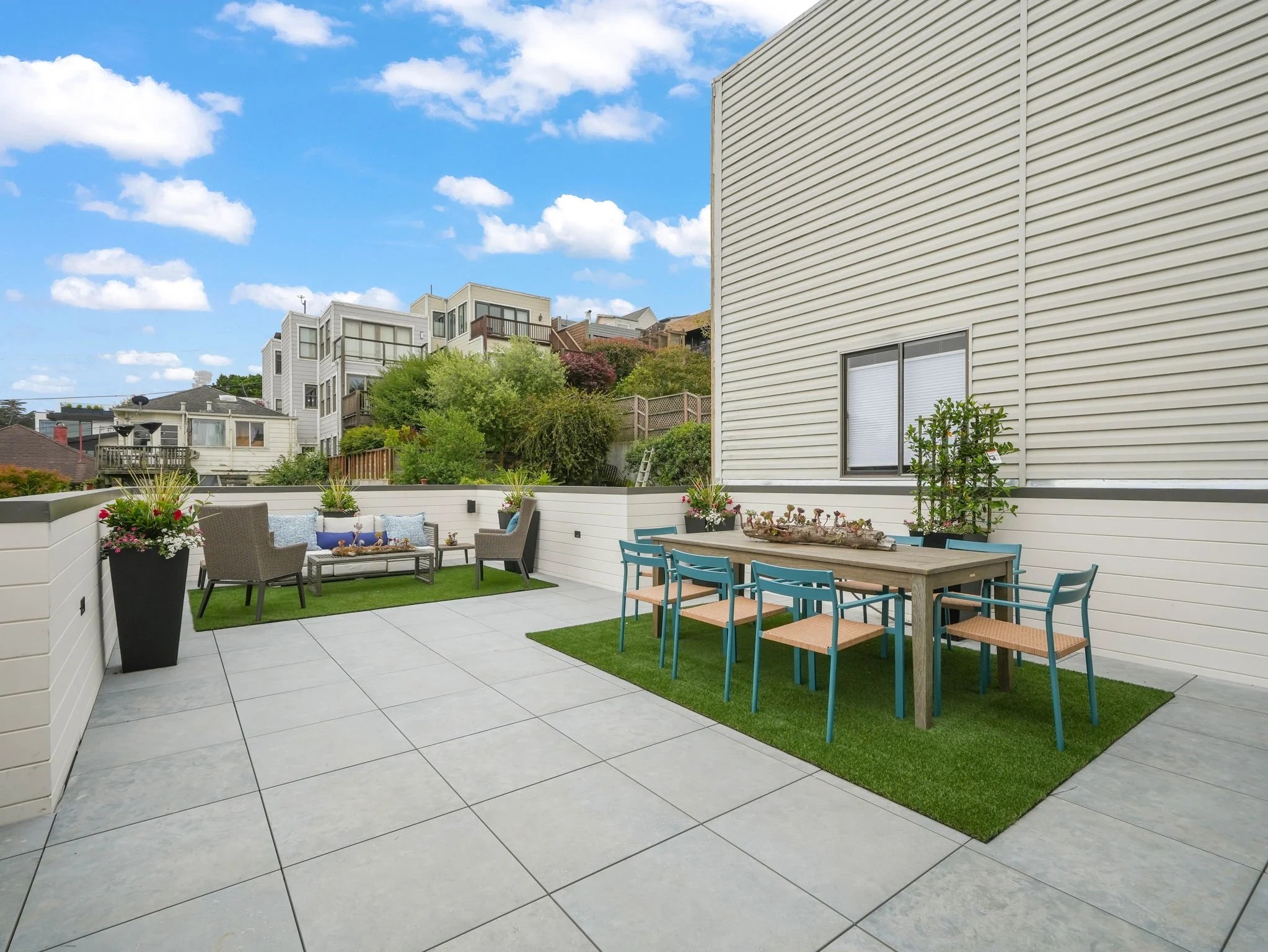 A rooftop patio with outdoor furniture, potted plants, a table with blue chairs, and a small artificial grass area, surrounded by a white fence, with neighboring houses and trees in the background under a partly cloudy sky.