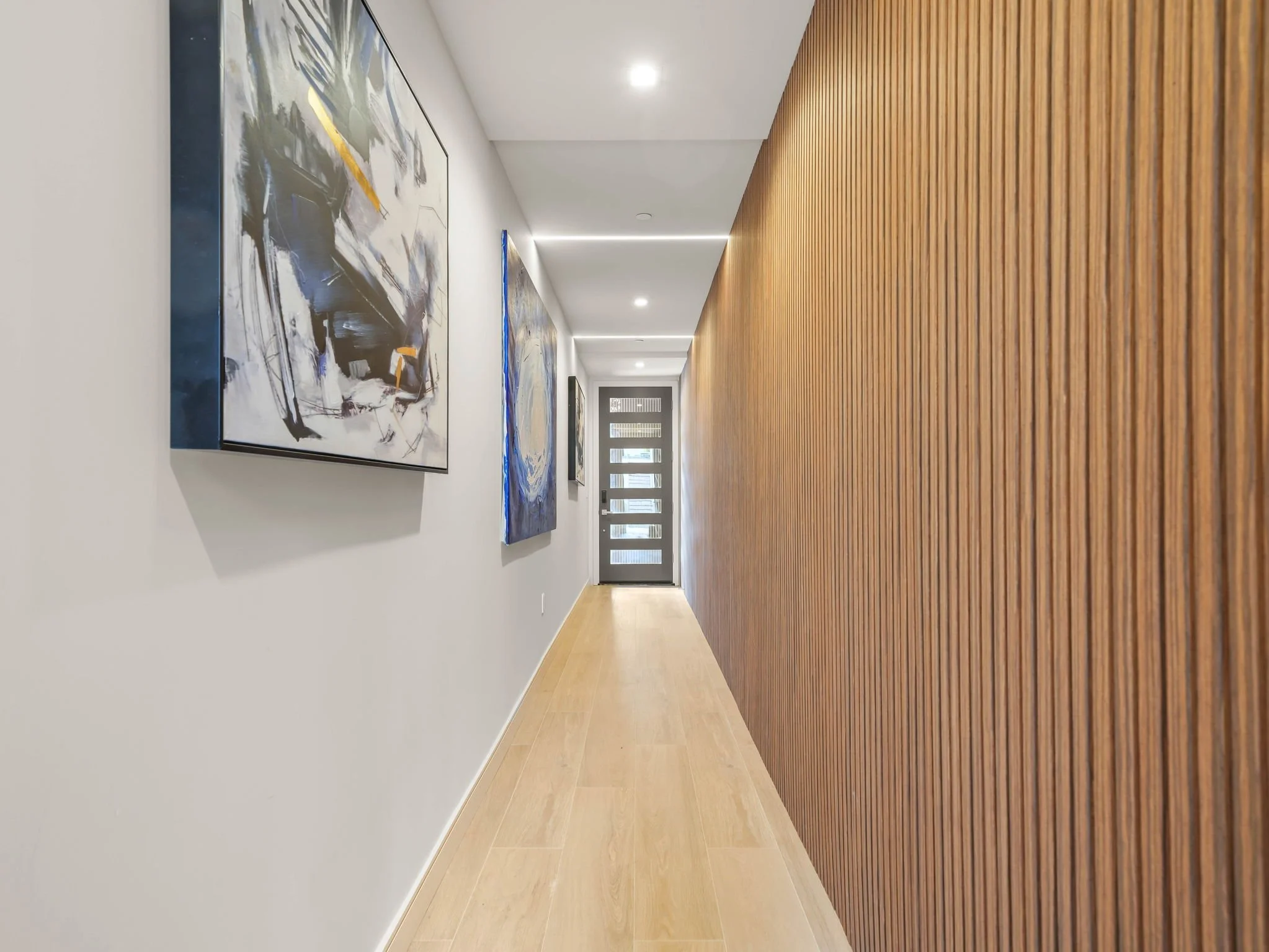 A modern hallway with a wooden slatted accent wall on the right and a plain white wall on the left, decorated with abstract paintings. The ceiling has recessed lighting and a door at the end of the hallway.
