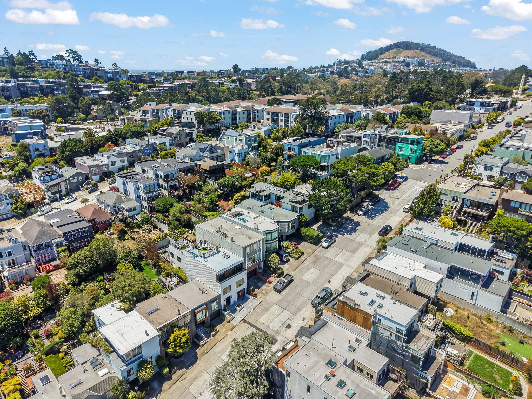 An aerial view of a residential neighborhood with houses, trees, and streets, with a hill in the background under a partly cloudy sky.