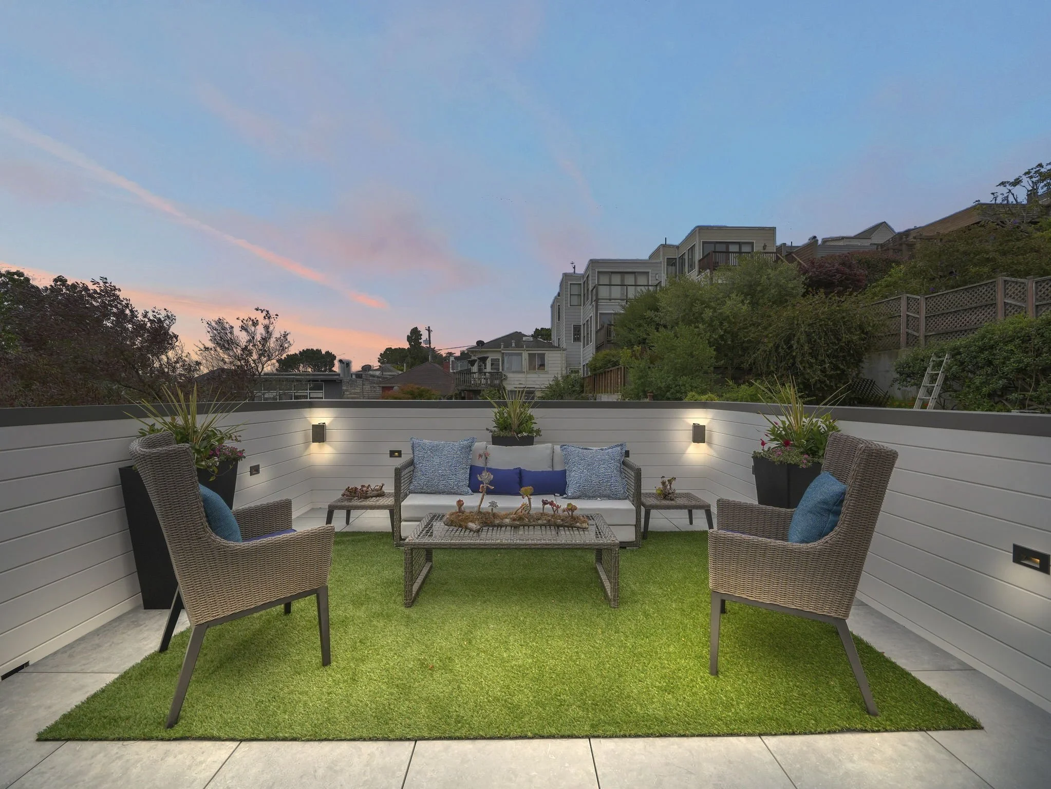 Outdoor patio with wicker chairs, a white planter with plants, and a grassy area surrounded by a white fence. In the background, there are residential houses and a hillside with trees and bushes, under a sky with pink and blue clouds at sunset.