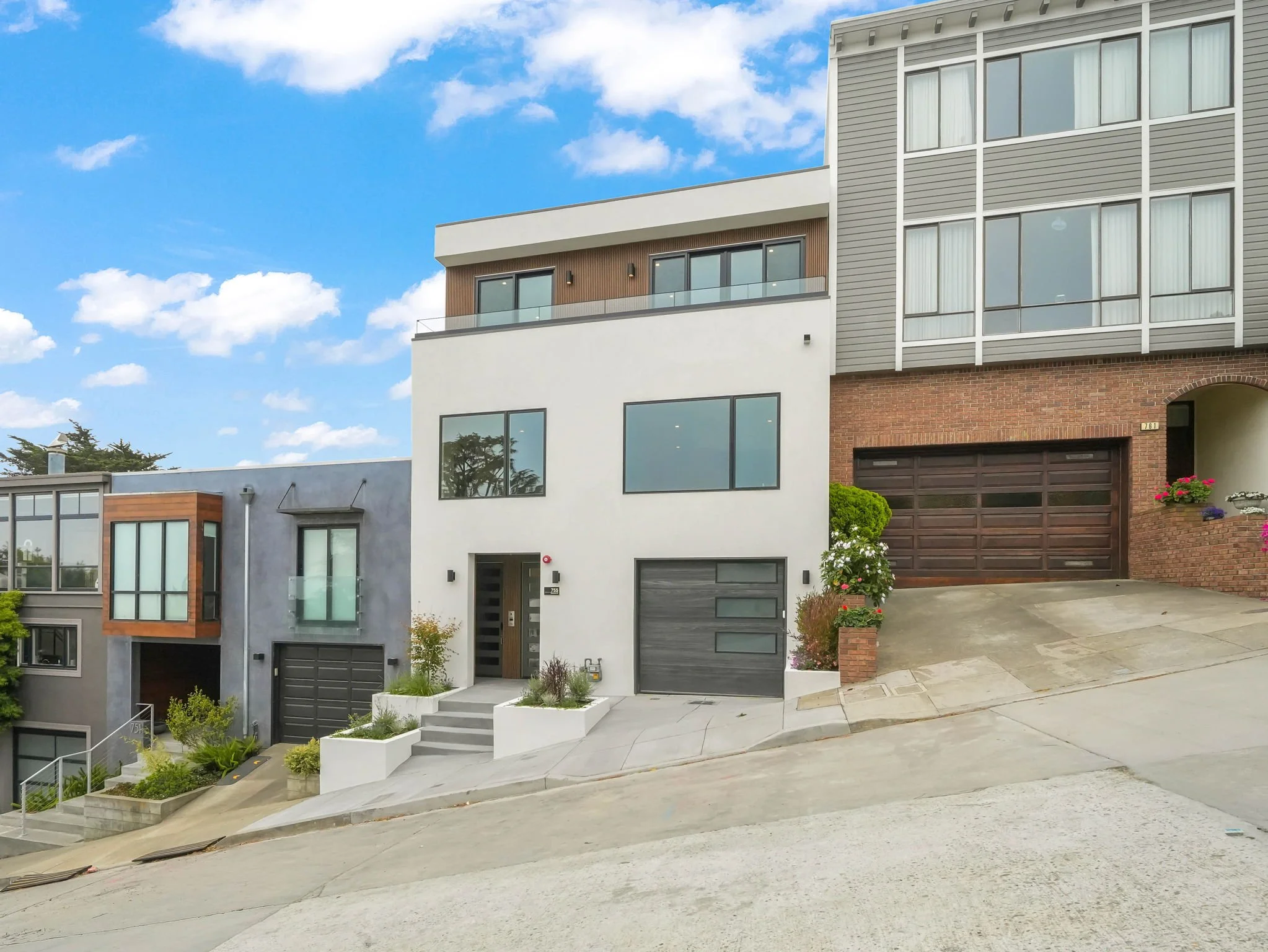 Modern multi-story residential buildings with large windows and garages, situated on a sloped street with stairs and small landscaped areas.