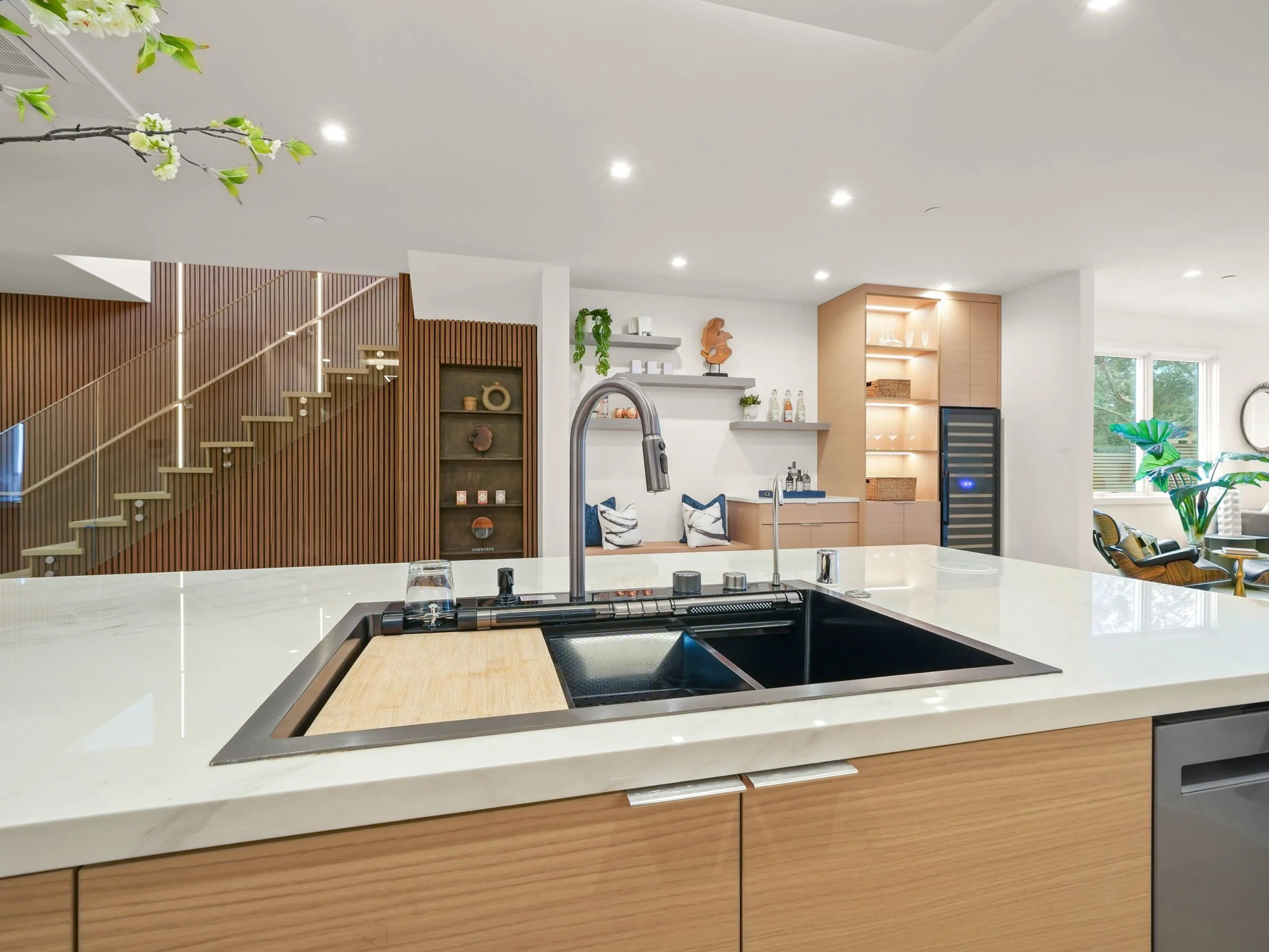 Modern kitchen with white countertop, stainless steel sink, and wooden cabinets, open to a living area with wall-mounted shelving, decorative items, and large window with greenery outside.