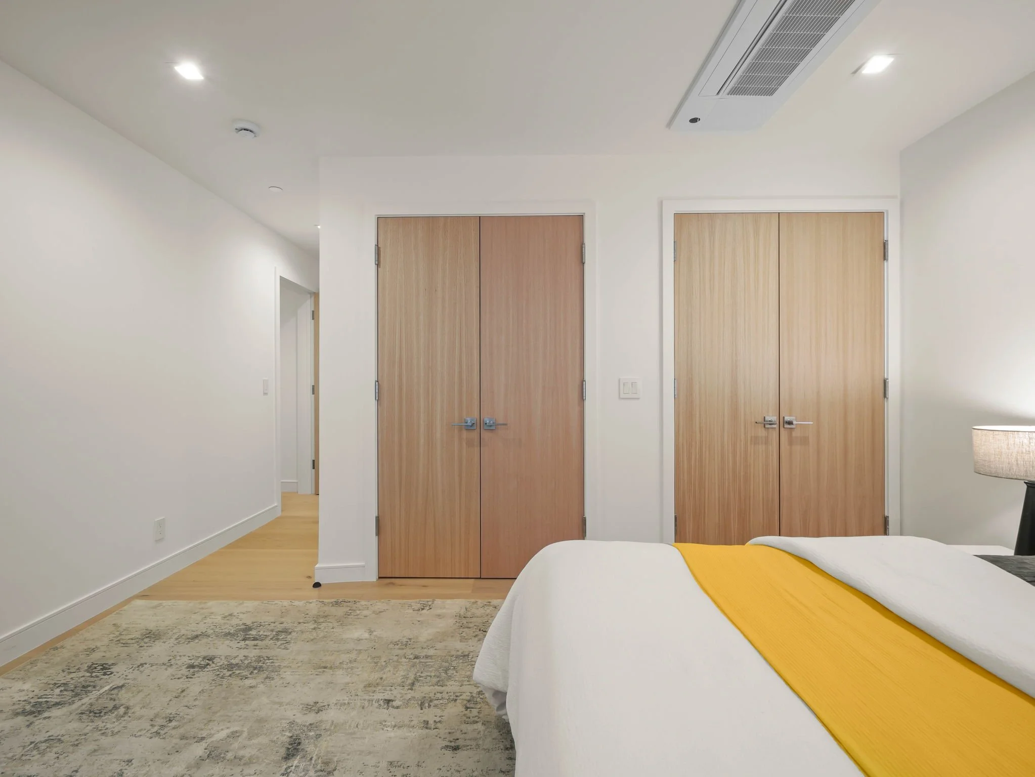 A bedroom with white walls, wooden doors, a rug, a bed with white bedding and yellow accents, and a lamp, with a ceiling air vent and lights.