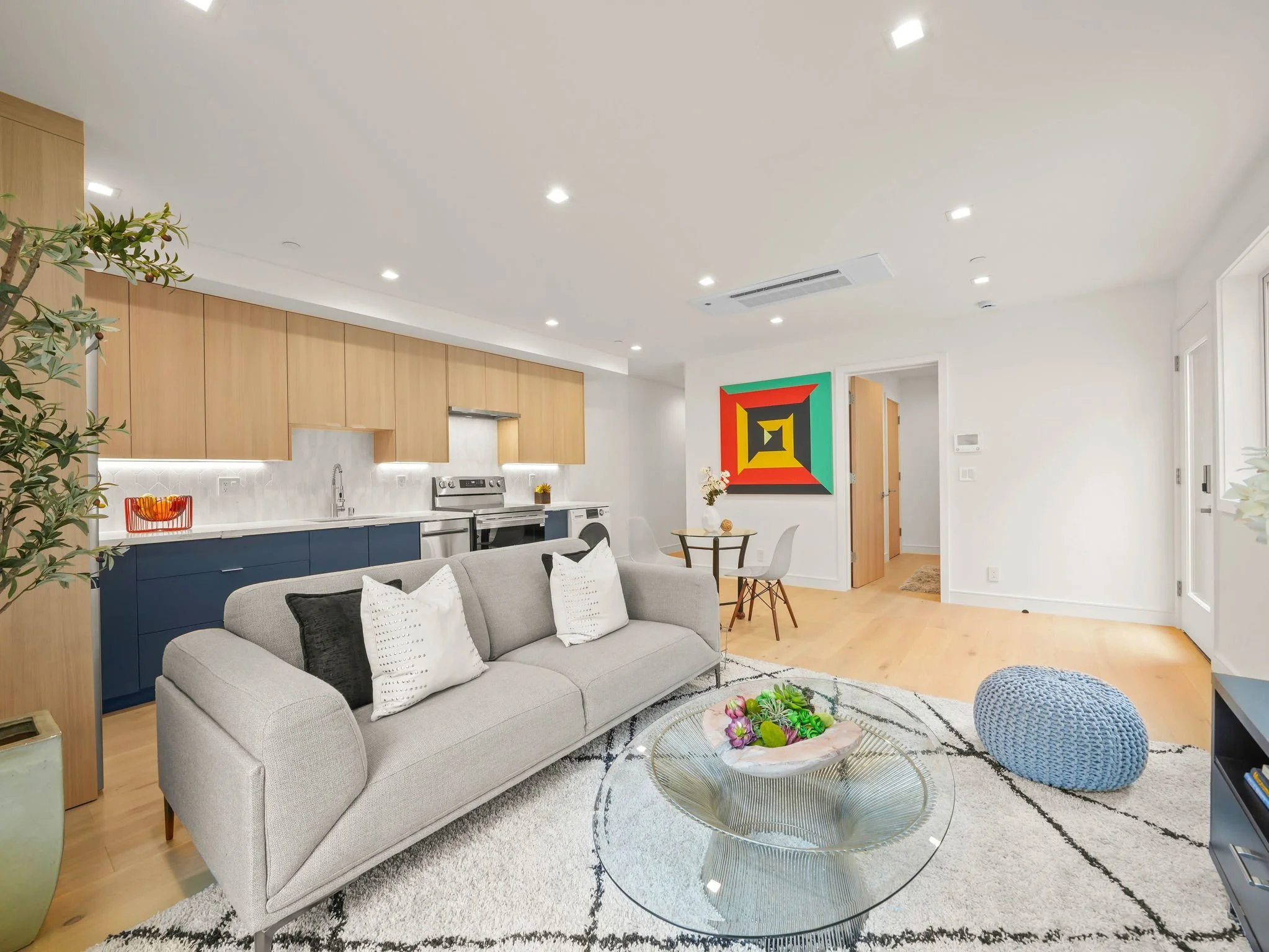 Modern living room and kitchen with beige sofa, colorful artwork, round glass coffee table, and wooden cabinets with white countertops.