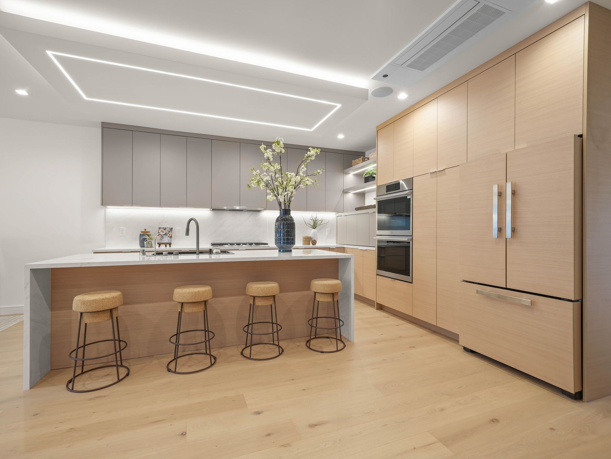 Modern kitchen with light wood cabinetry, white marble backsplash, white countertop, and a kitchen island with three beige stools. The ceiling has decorative LED strip lighting and recessed lights. A vase with white flowers is on the island.