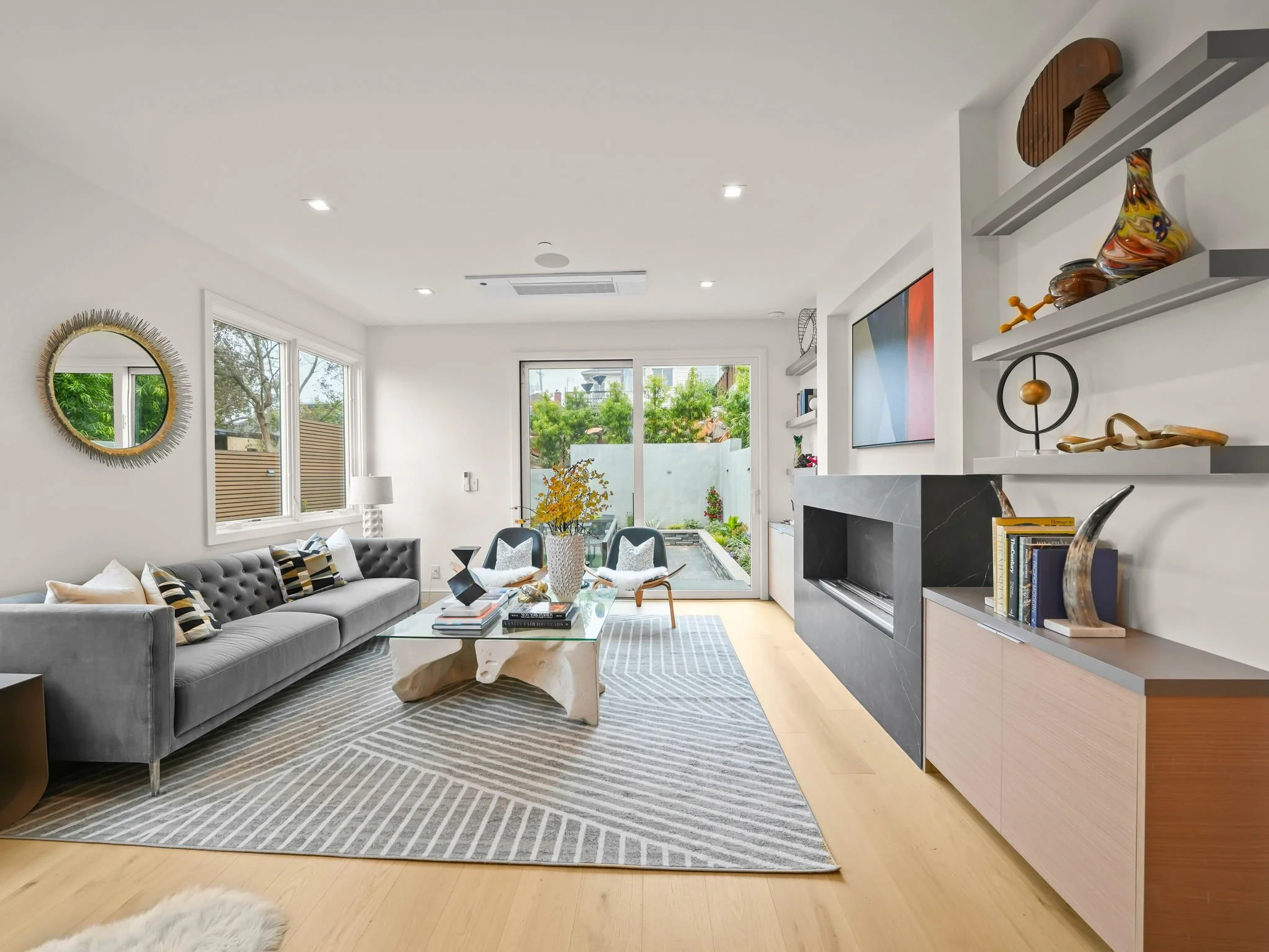 Bright living room with a gray sofa, patterned pillows, glass coffee table, and a large window with a view of trees and outdoor space.