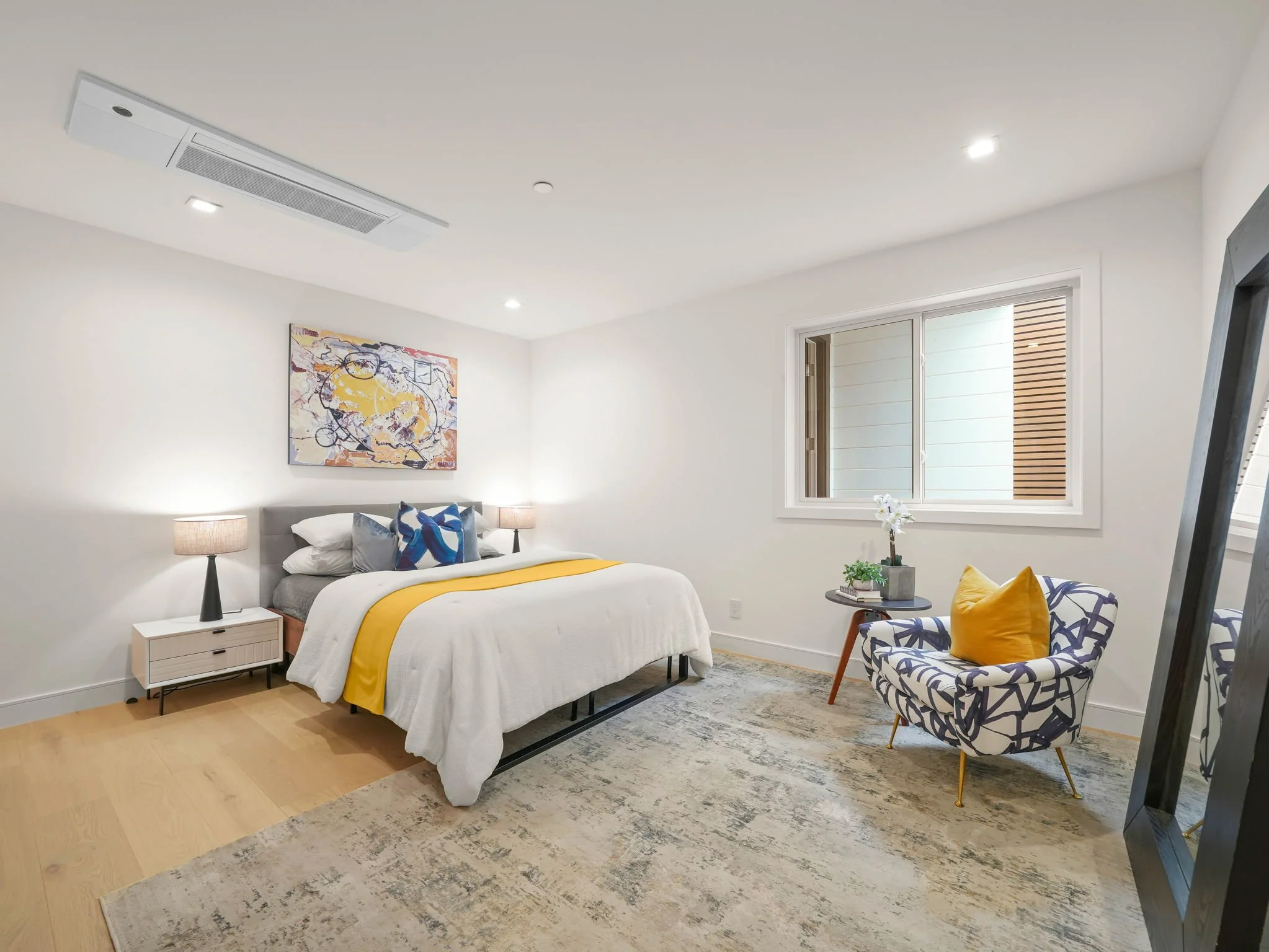 A bedroom with a bed, nightstands, lamps, a colorful piece of abstract art, a window, an armchair with a yellow pillow, and a decorative rug.