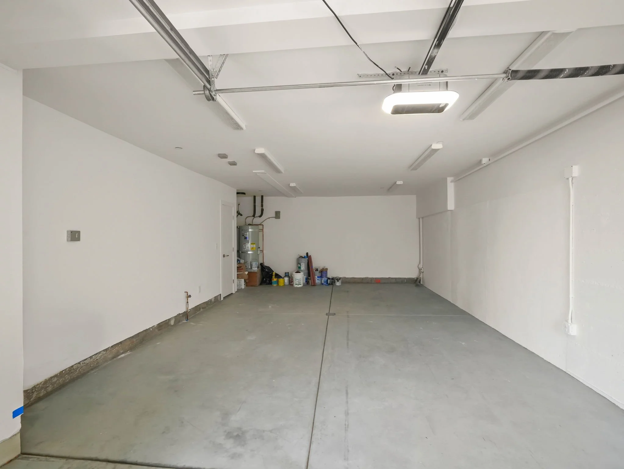 Empty garage with white walls, unfinished concrete floor, and a ceiling with exposed wiring and lighting fixtures. Construction supplies and equipment are stored along the back wall.