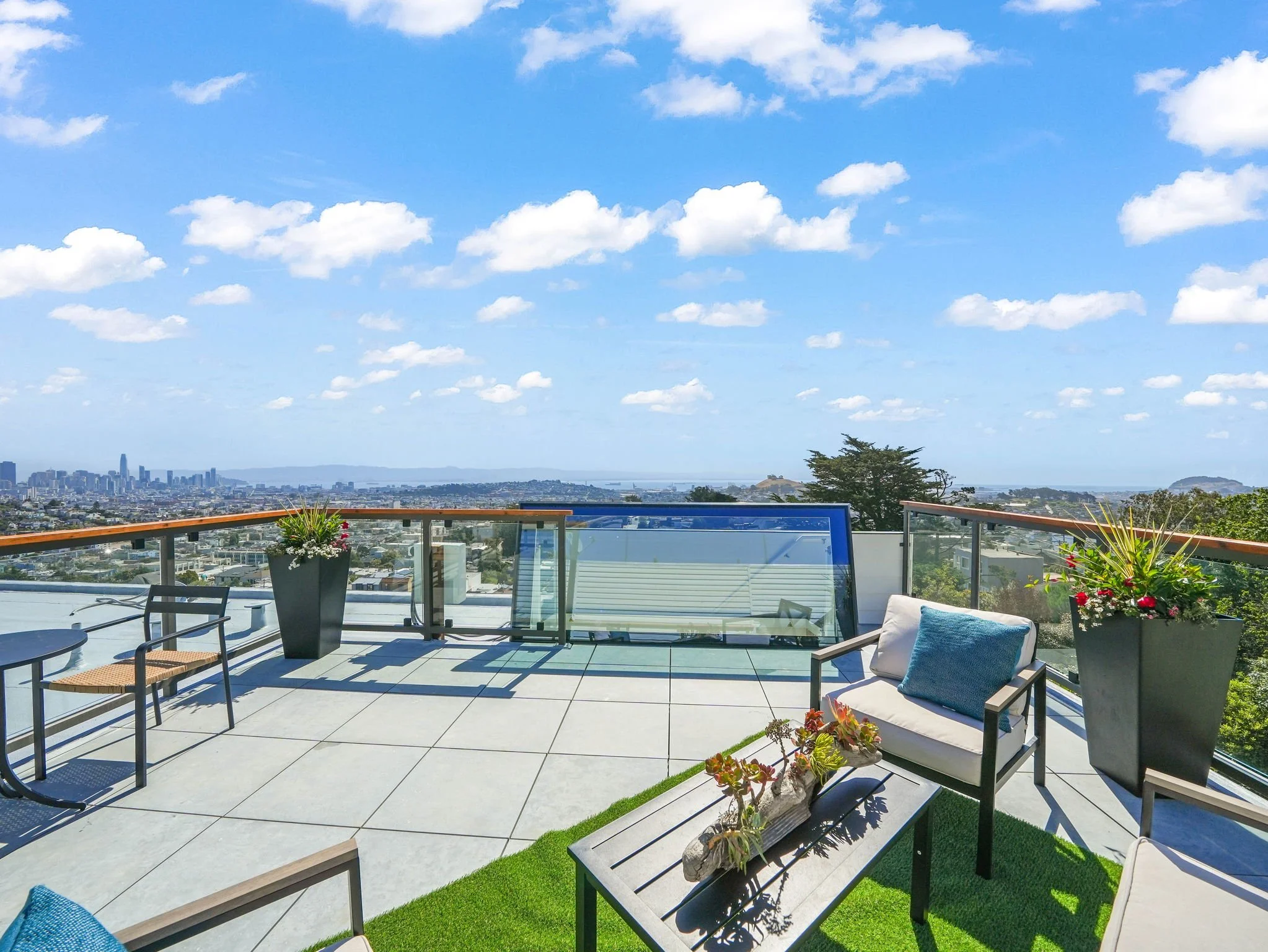 A rooftop terrace with outdoor furniture, including tables, chairs, and a cushioned lounge chair, surrounded by potted plants and overlooking a city skyline with skyscrapers, a bay, and a partly cloudy sky.