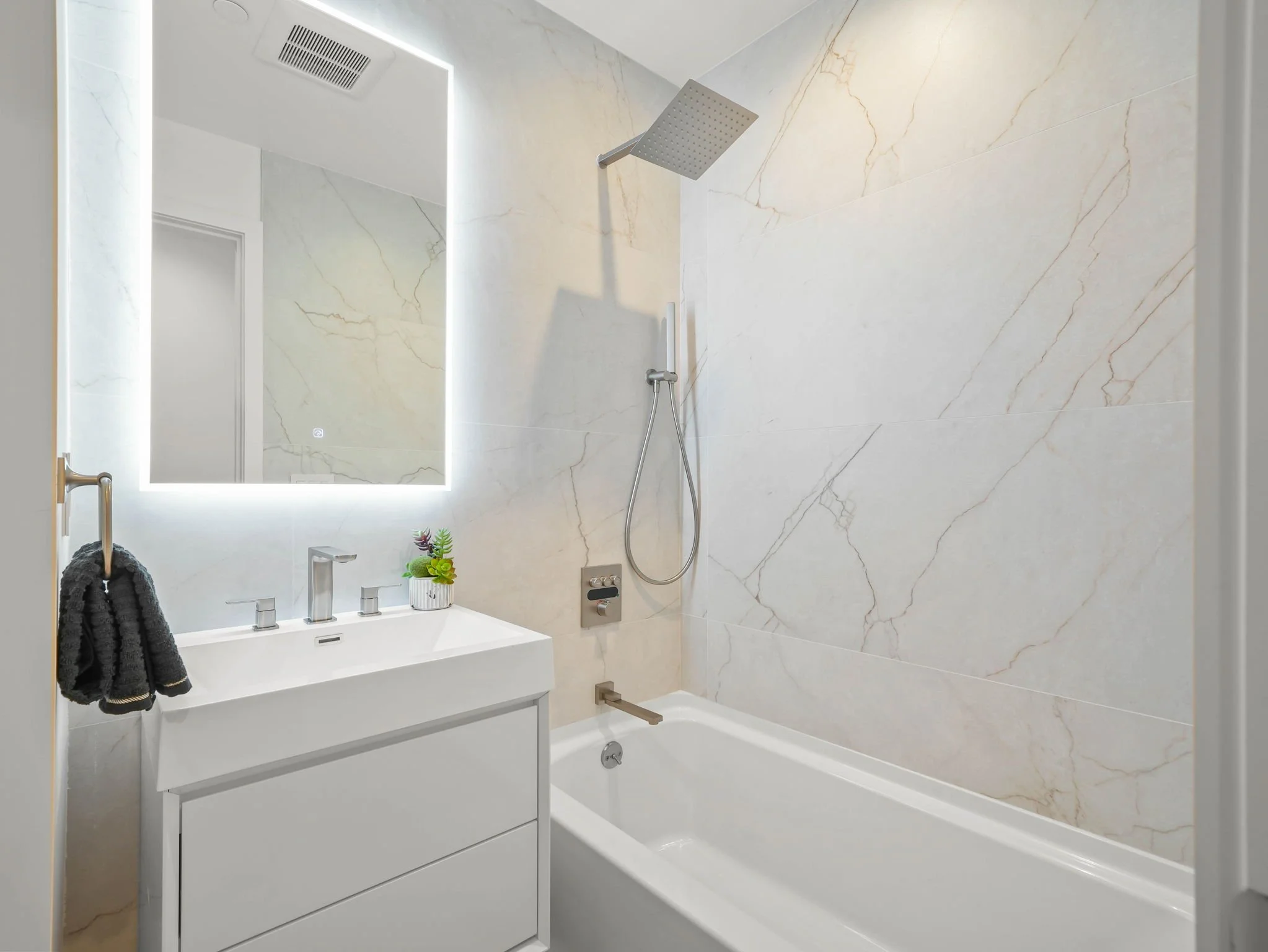 Modern bathroom with white vanity, large wall mirror with backlit lighting, marble wall tiles, rainfall showerhead, and a small plant on the vanity.