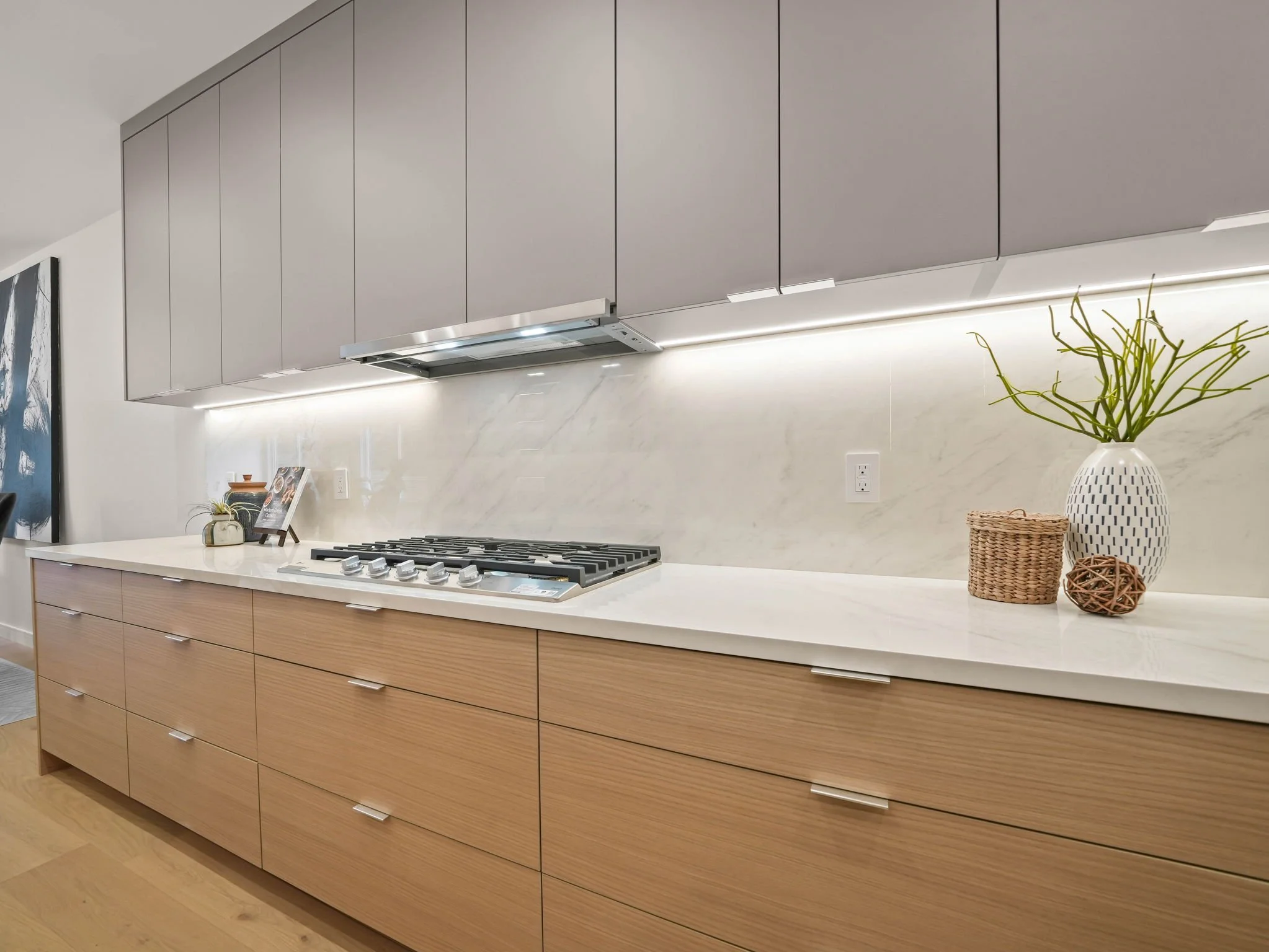 Modern kitchen with a light wood island, gray upper cabinets, white marble backsplash, and a stove with four burners. Decor includes a white vase with green plants and small baskets on the countertop.