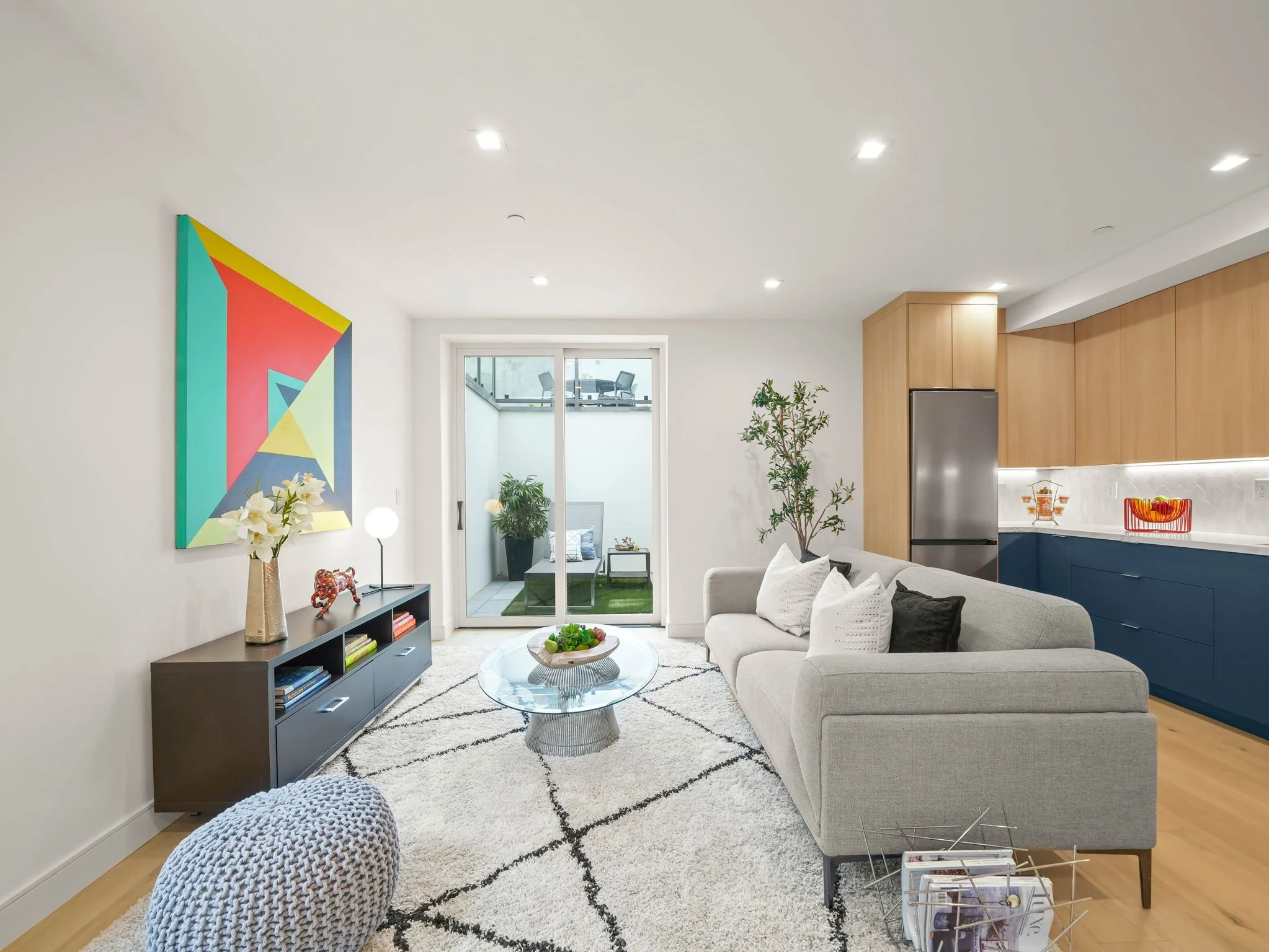 Living room with a beige sofa, a black console table with books and decorations, a round glass coffee table, a patterned white rug, green plants, a colorful geometric wall art, and a sliding glass door leading to an outdoor seating area.