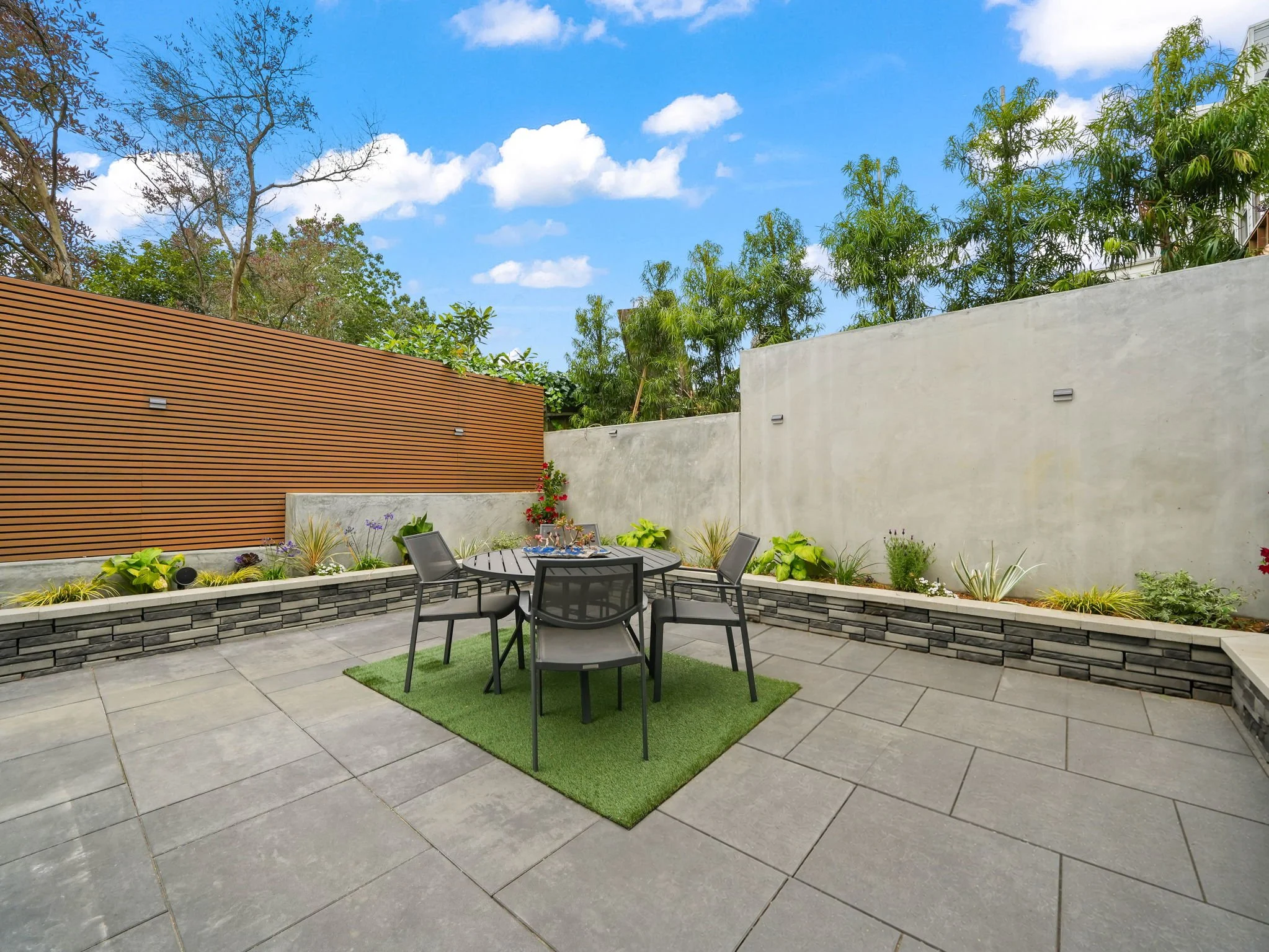 A modern outdoor patio with gray tiled flooring, a circular black metal table, and four matching chairs on a small green rug. The patio is surrounded by a low stone wall with decorative plants and flowers. Tall concrete and wooden privacy walls with 