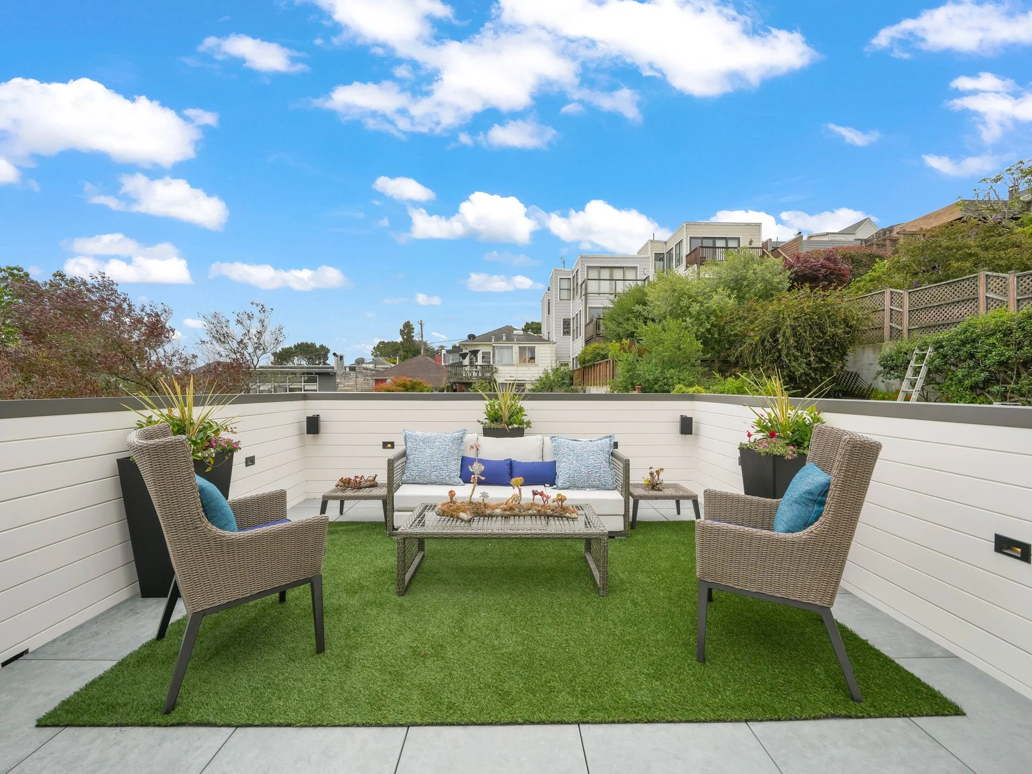 A cozy rooftop patio with patio furniture, potted plants, and a view of residential buildings and a partly cloudy blue sky.