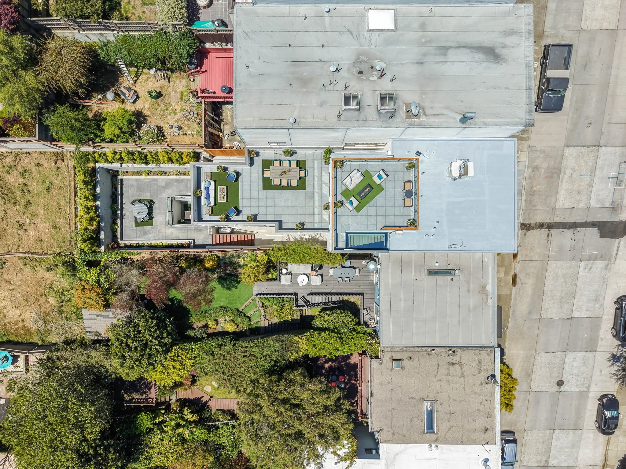 An aerial view of a building with a flat gray roof, surrounded by a parking lot and greenery, including trees and shrubs, with longer shadows cast by objects suggesting sunlight.