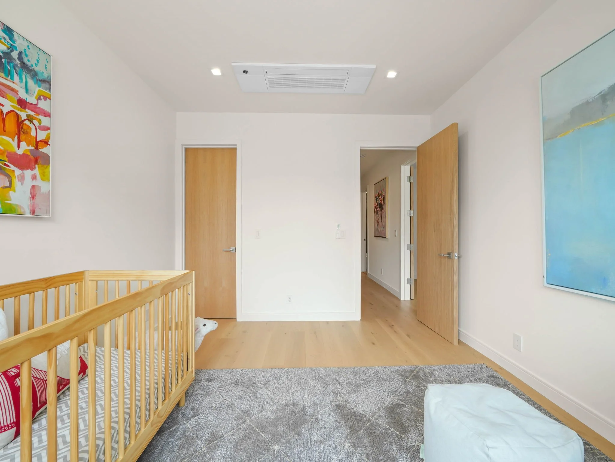 A nursery room with white walls, light wood flooring, a wooden crib, colorful abstract paintings on the walls, two closed wooden doors, and an open door leading to another room with a framed artwork on the wall.
