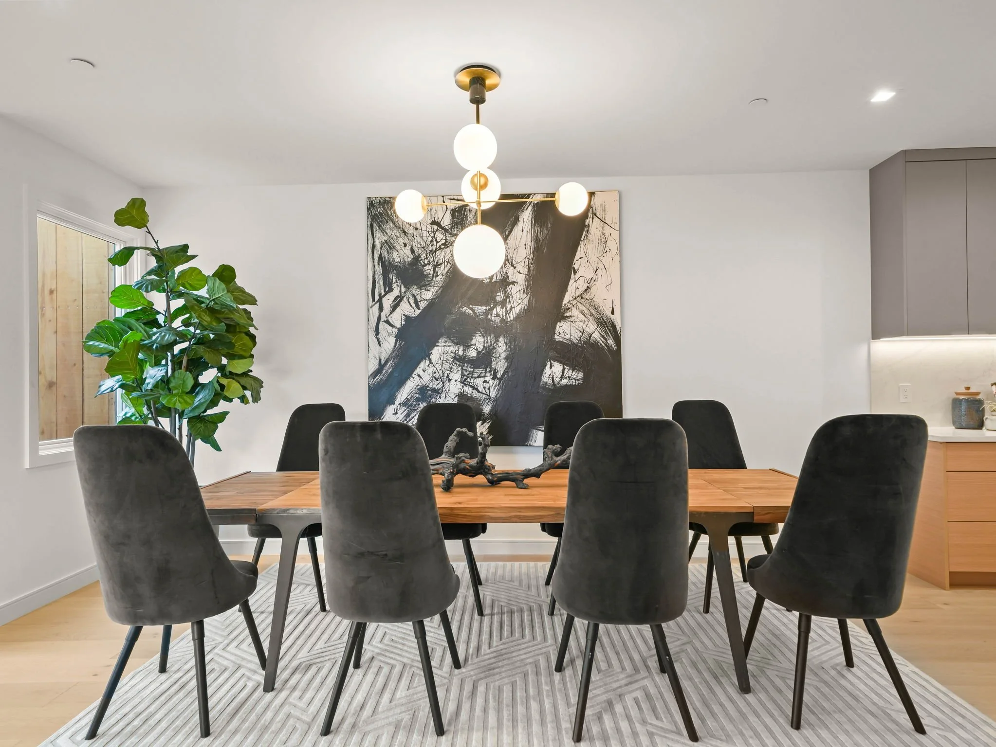 A contemporary dining room featuring a wooden table surrounded by six gray velvet chairs, a large abstract black and white painting on the wall, a green potted plant near a window, and modern lighting fixtures hanging from a white ceiling.