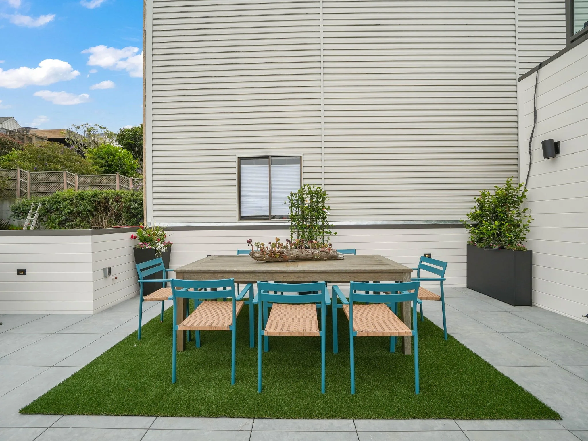 A backyard patio with a rectangular wooden dining table surrounded by blue chairs with woven seats, set on a green artificial grass rug. The patio is enclosed by white siding walls, with potted plants and flowers along the perimeter. An outdoor black