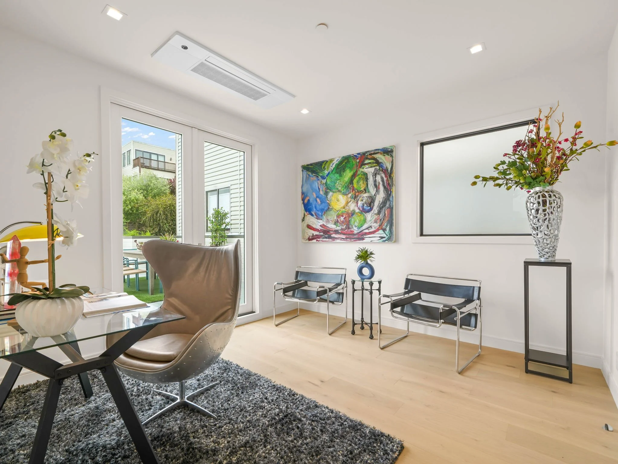 Bright modern room with large windows, white walls, a colorful abstract painting, potted plants, and modern black chairs.