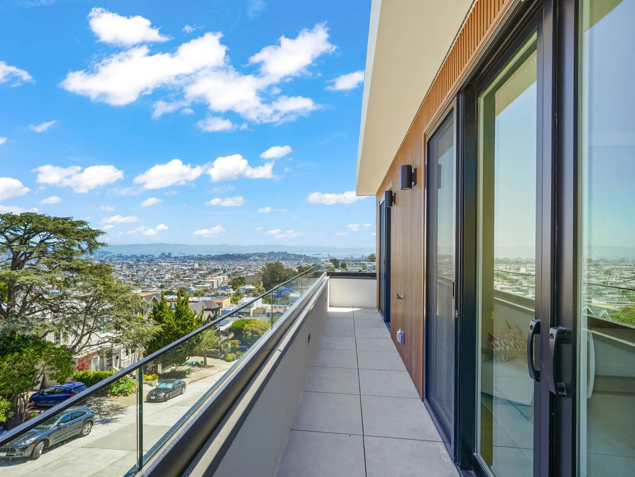 A modern balcony with glass railing overlooking a cityscape, with a view of trees, rooftops, and distant hills under a blue sky with white clouds.
