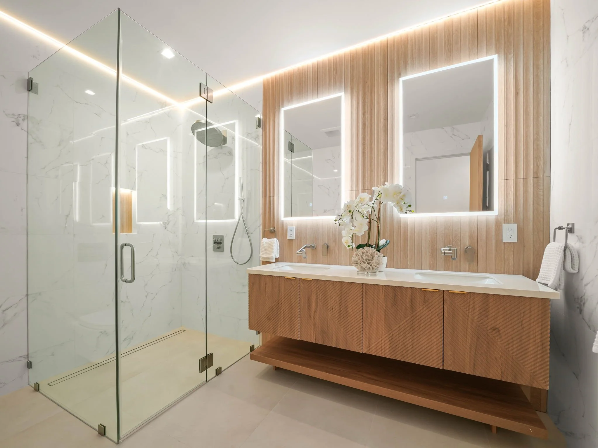 Modern bathroom with a glass shower enclosure, white marble walls, and a wooden vanity with a large mirror surrounded by LED lighting.