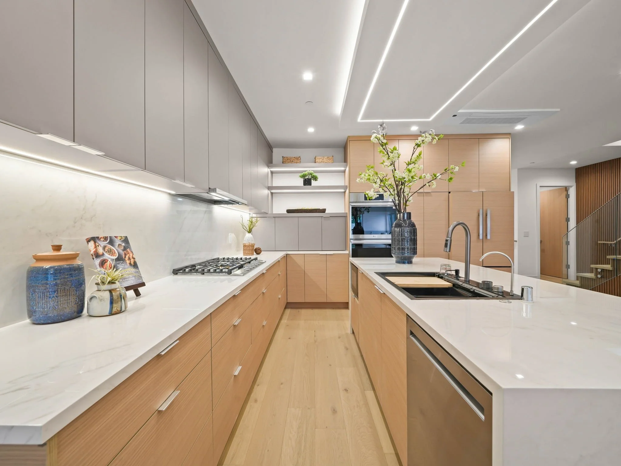 Modern kitchen with white countertops, light wood cabinets, and decorative vases on the counter. Includes a stove, sink, and large flowering plant as decor.