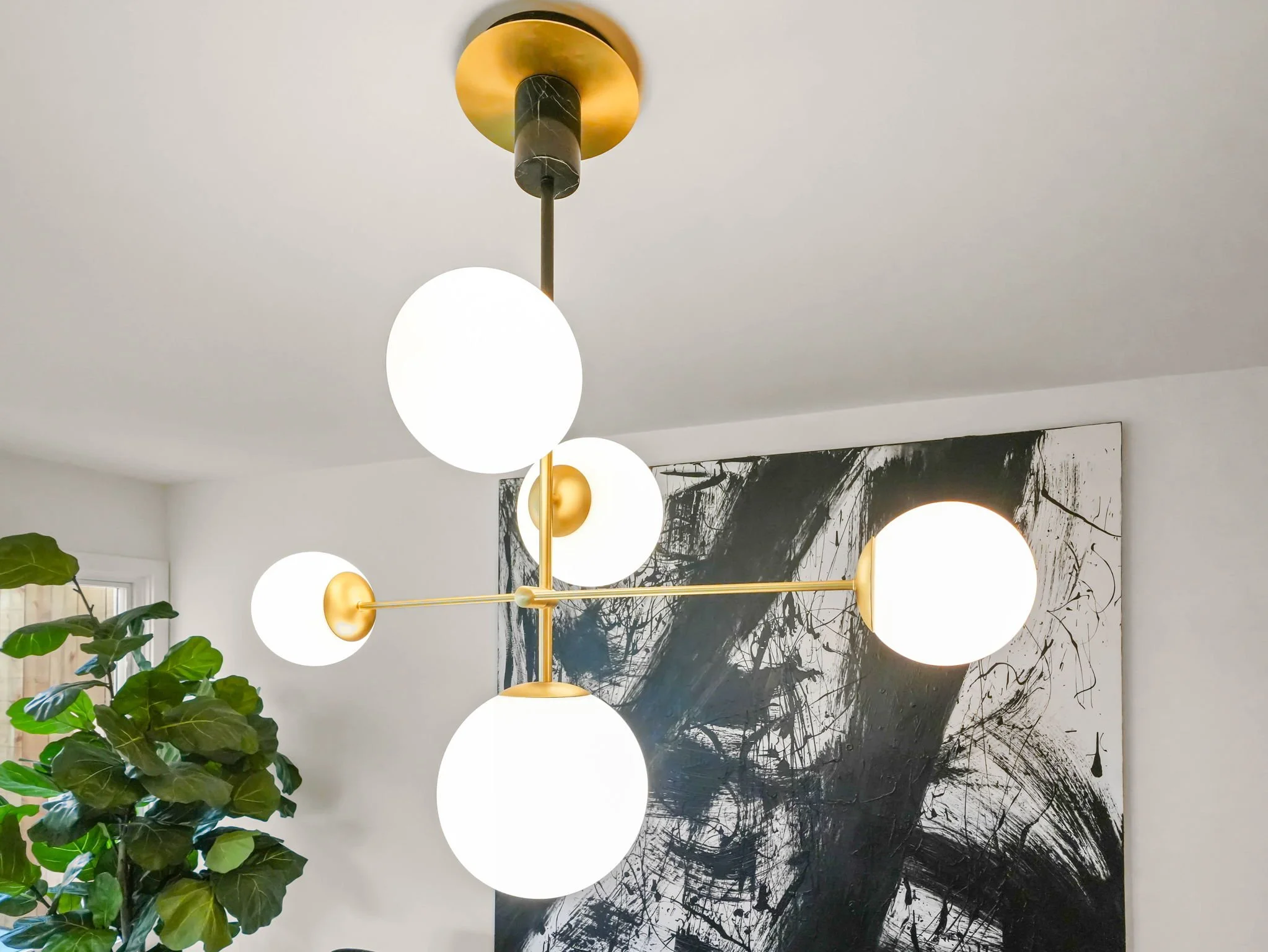 Modern chandelier with six round white globes hanging from a gold fixture, positioned above a black and white abstract painting, with a green leafy plant nearby.