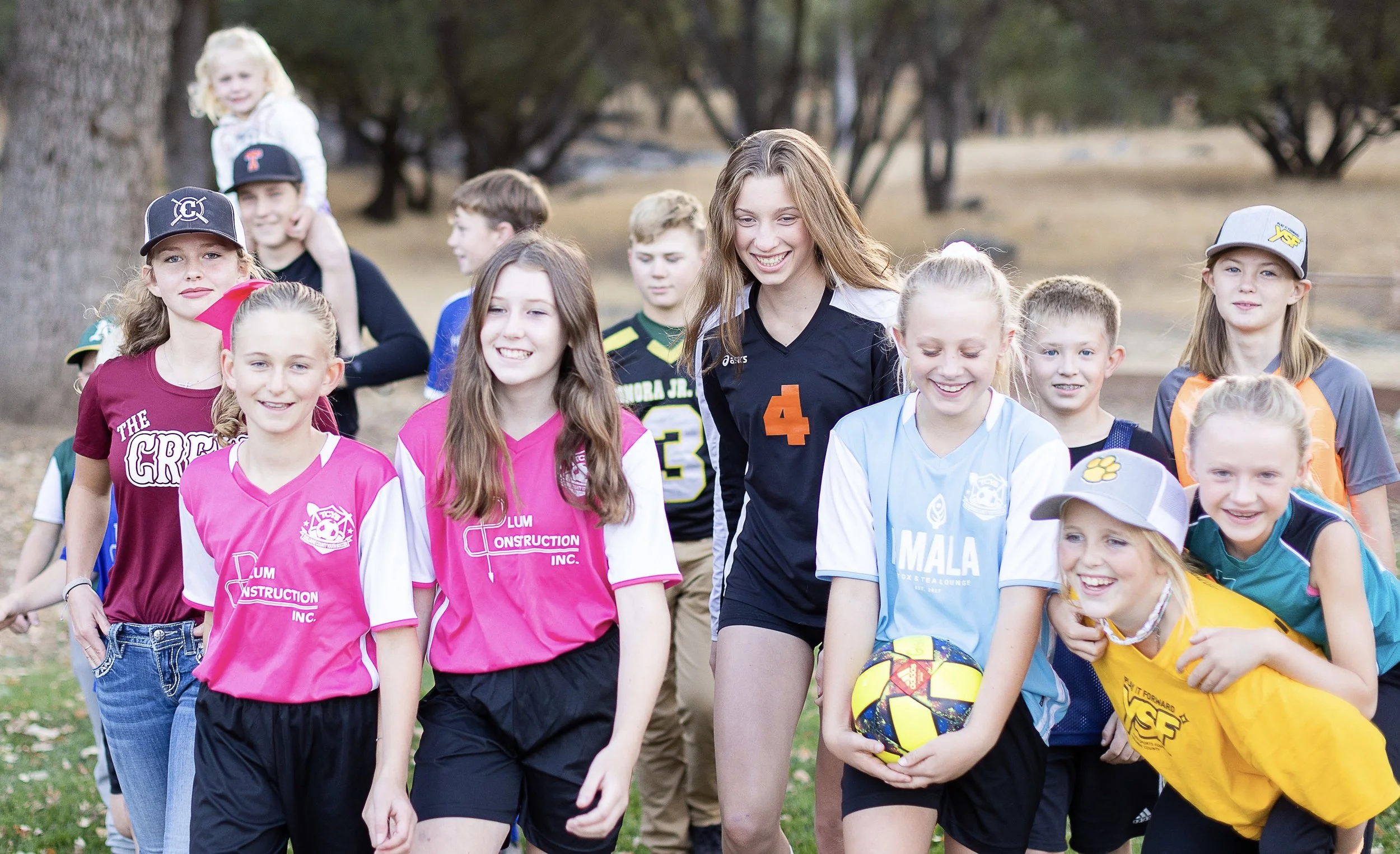 Group of children and a teenage girl walking outdoors in a park, smiling and wearing sports jerseys and caps, with a tree in the background.